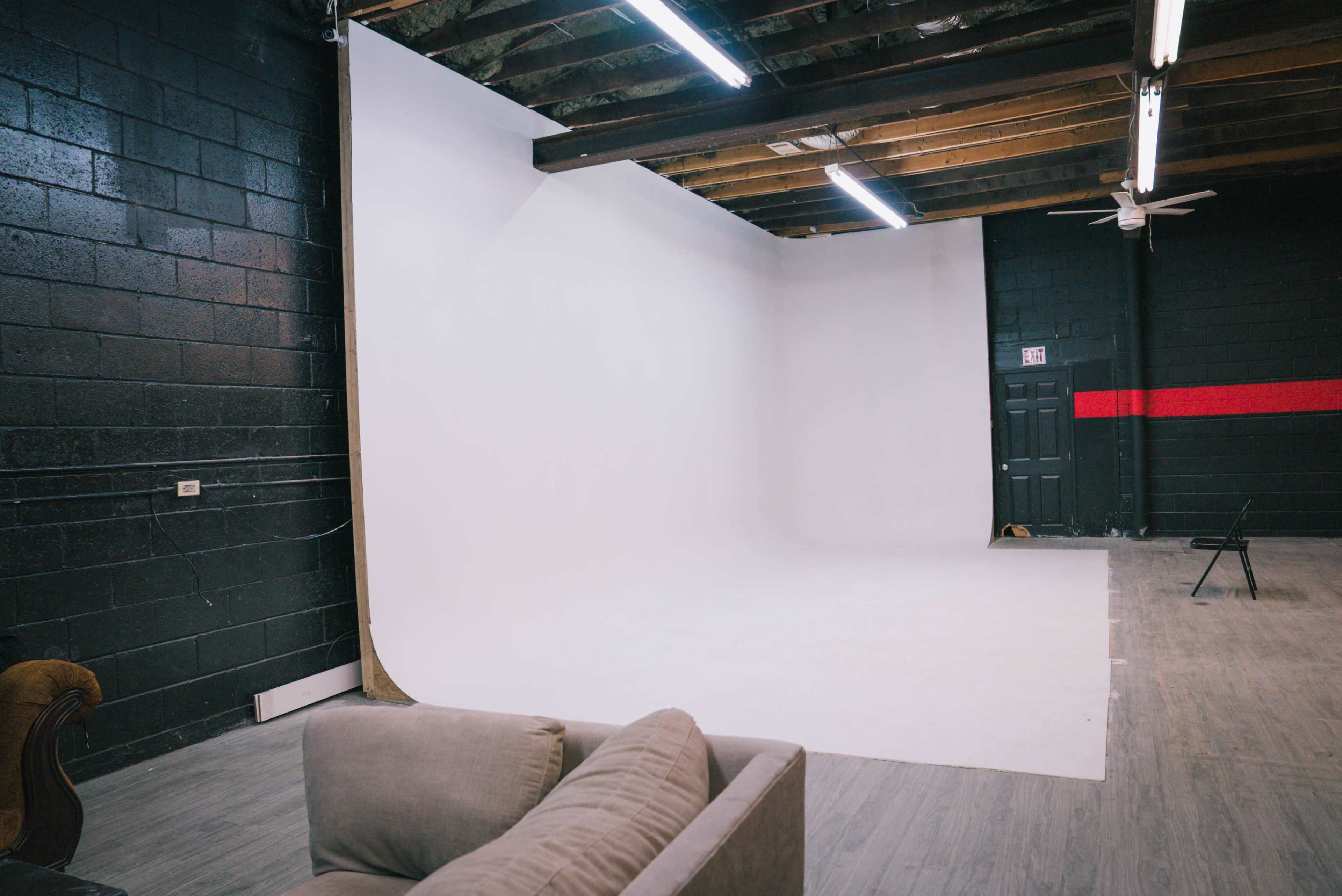 The image shows a spacious studio with a white backdrop, black walls, exposed wooden beams, and a simple couch in the foreground.