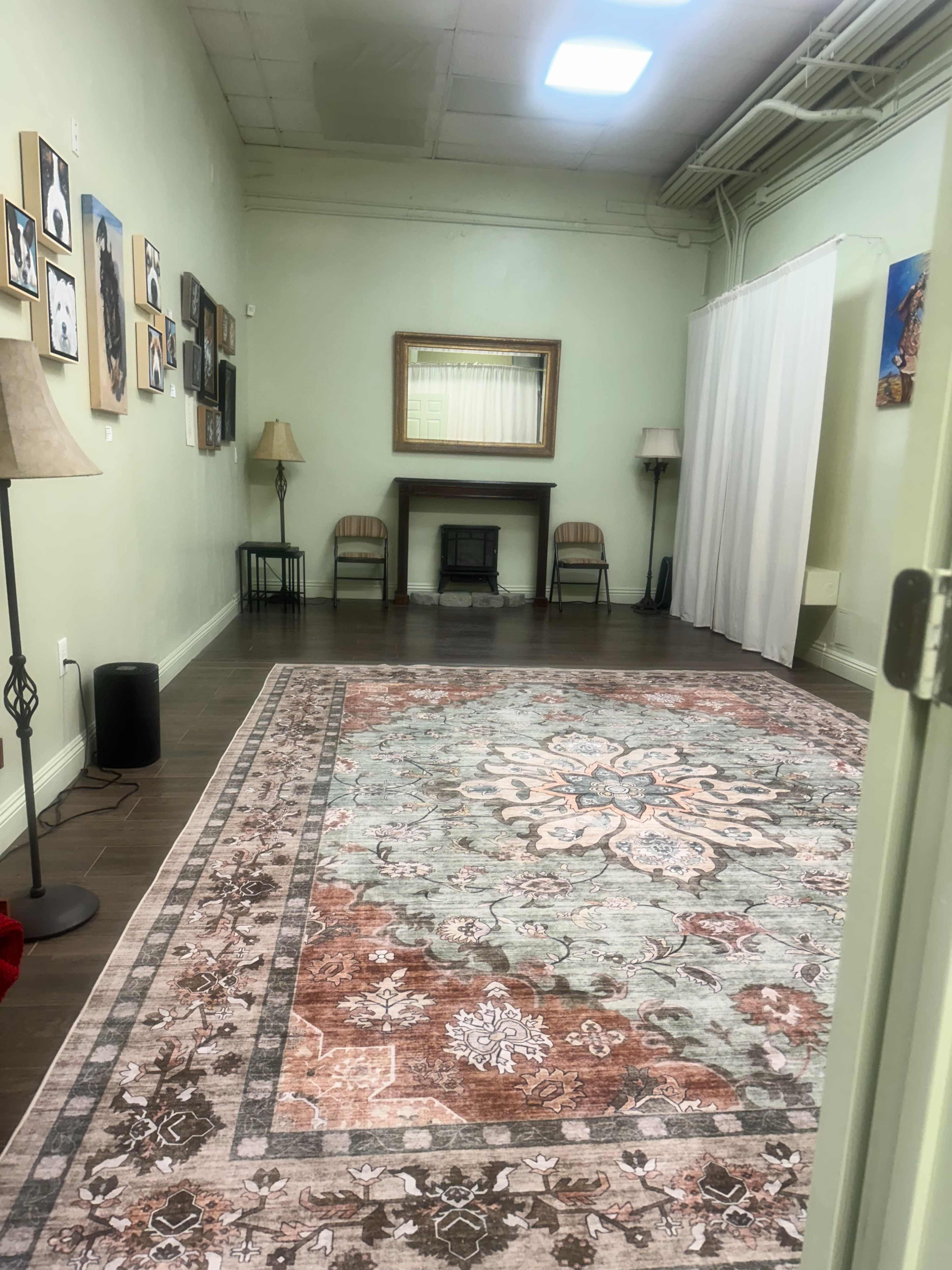 The image shows a spacious room with a patterned area rug, two lamps, artwork on the walls, and a small fireplace with seating nearby.
