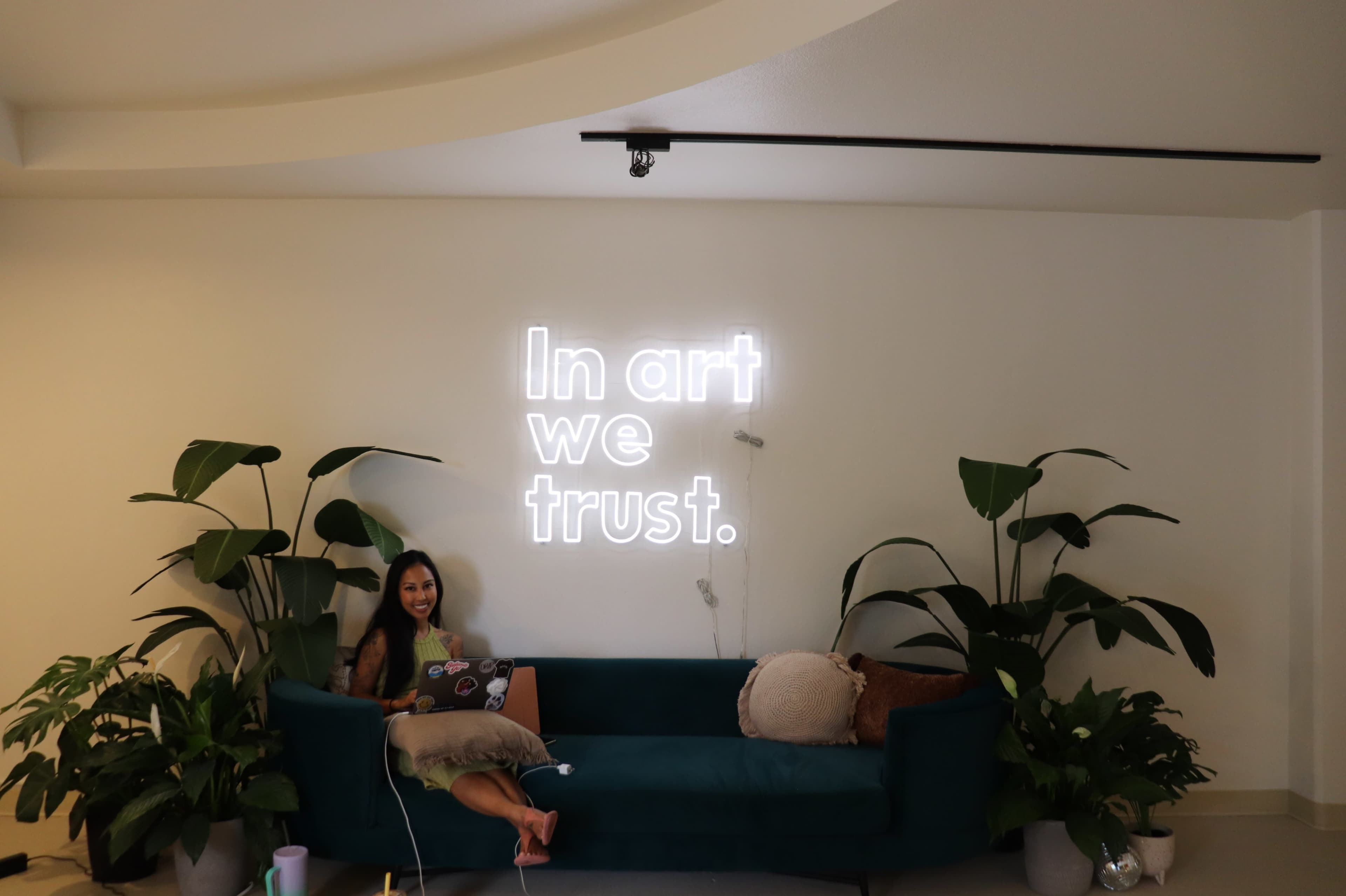 A person sits on a teal couch with plants, working on a laptop in front of a neon sign that says "In art we trust."