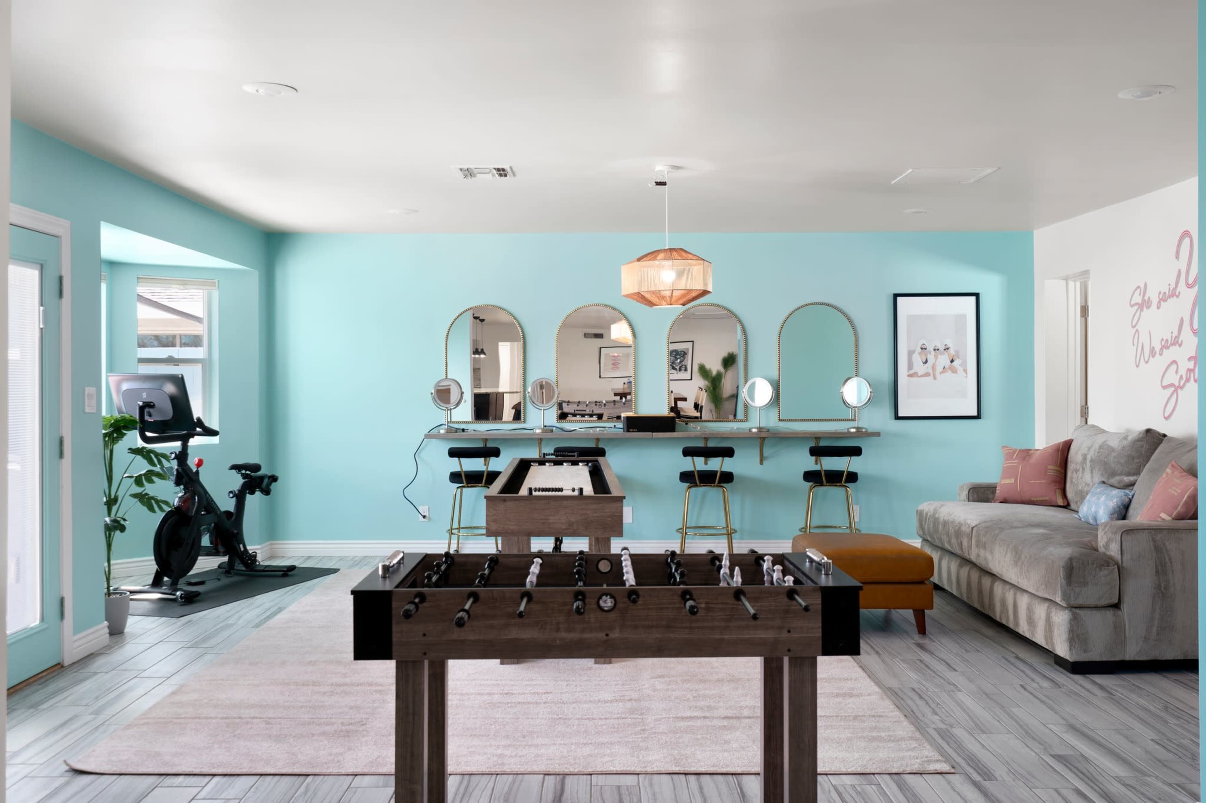 A game room features a foosball table in the foreground, a bar with stools in the background, and a seating area with a couch and decorative elements.