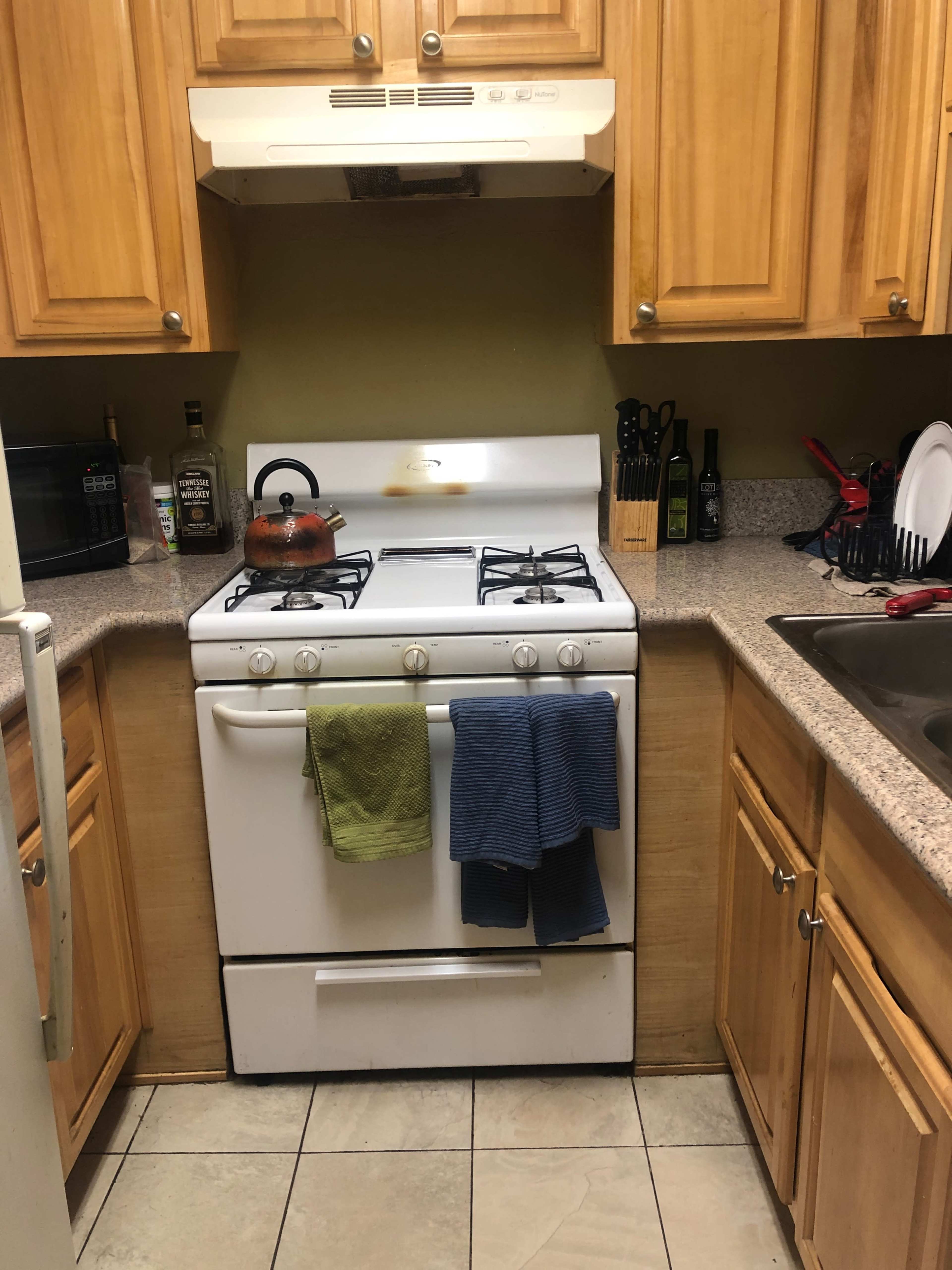 A small kitchen with wooden cabinets, a white gas stove, a sink, and various kitchen utensils arranged on the counter.