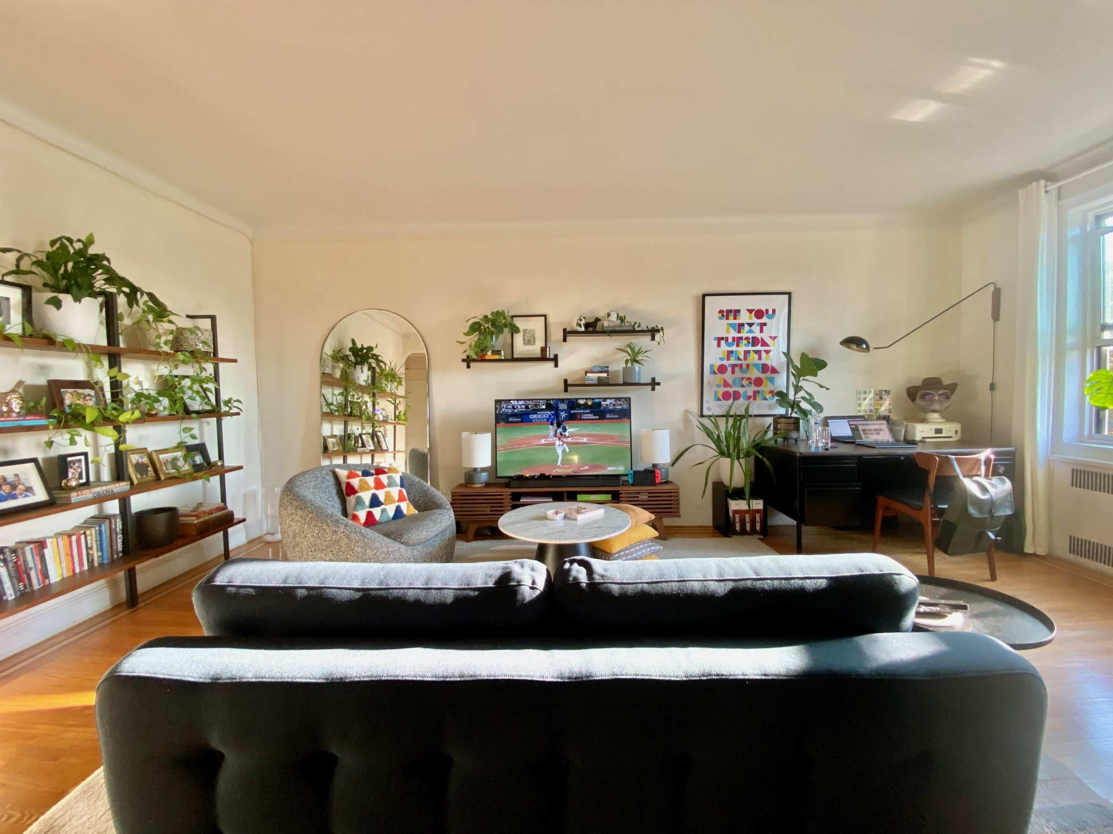 A cozy living room features a sofa, a coffee table, a television displaying a sports game, and shelves filled with plants and books.