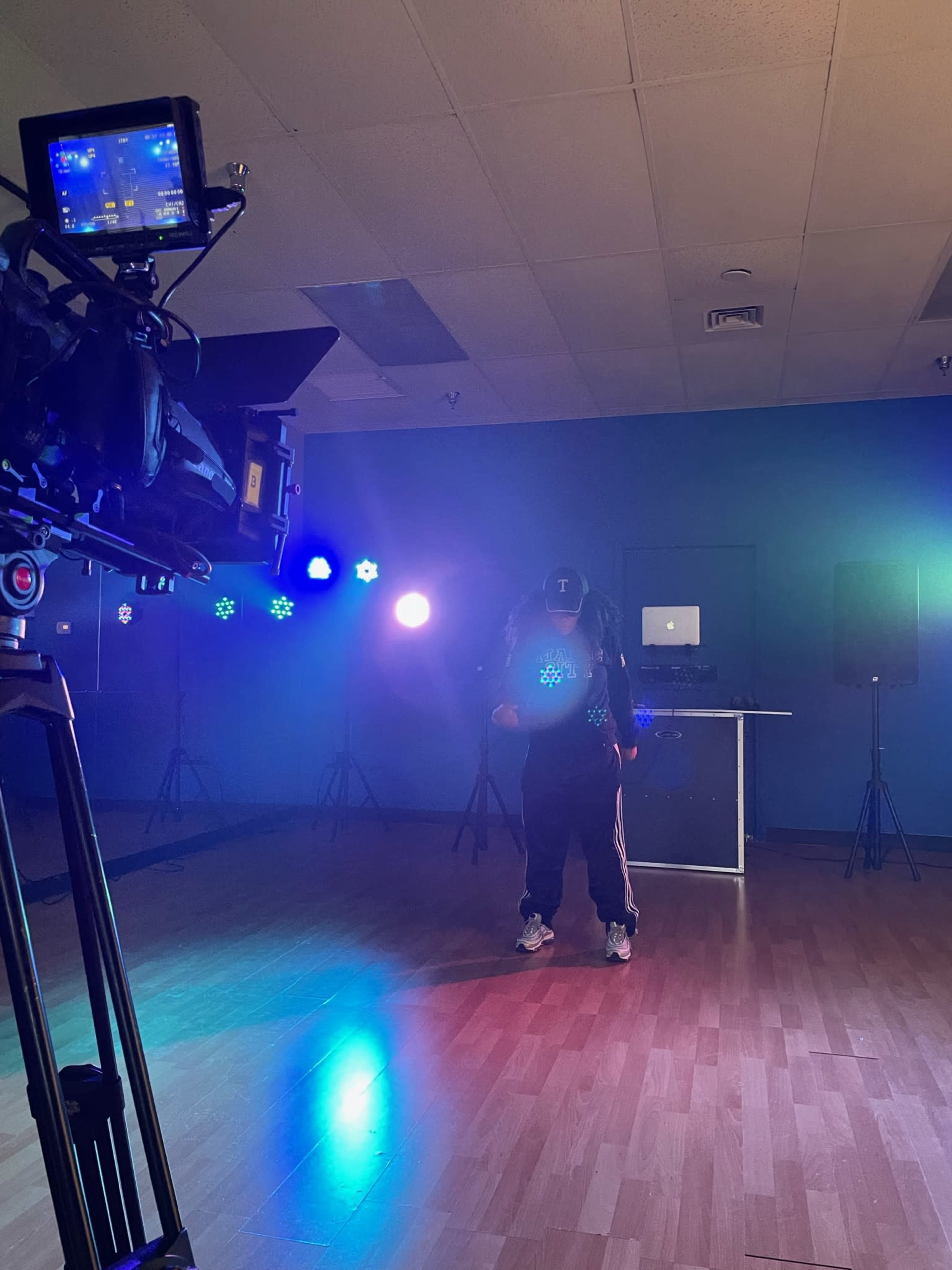 A person in athletic wear dances in a lighted studio space with a camera and DJ equipment in the background.