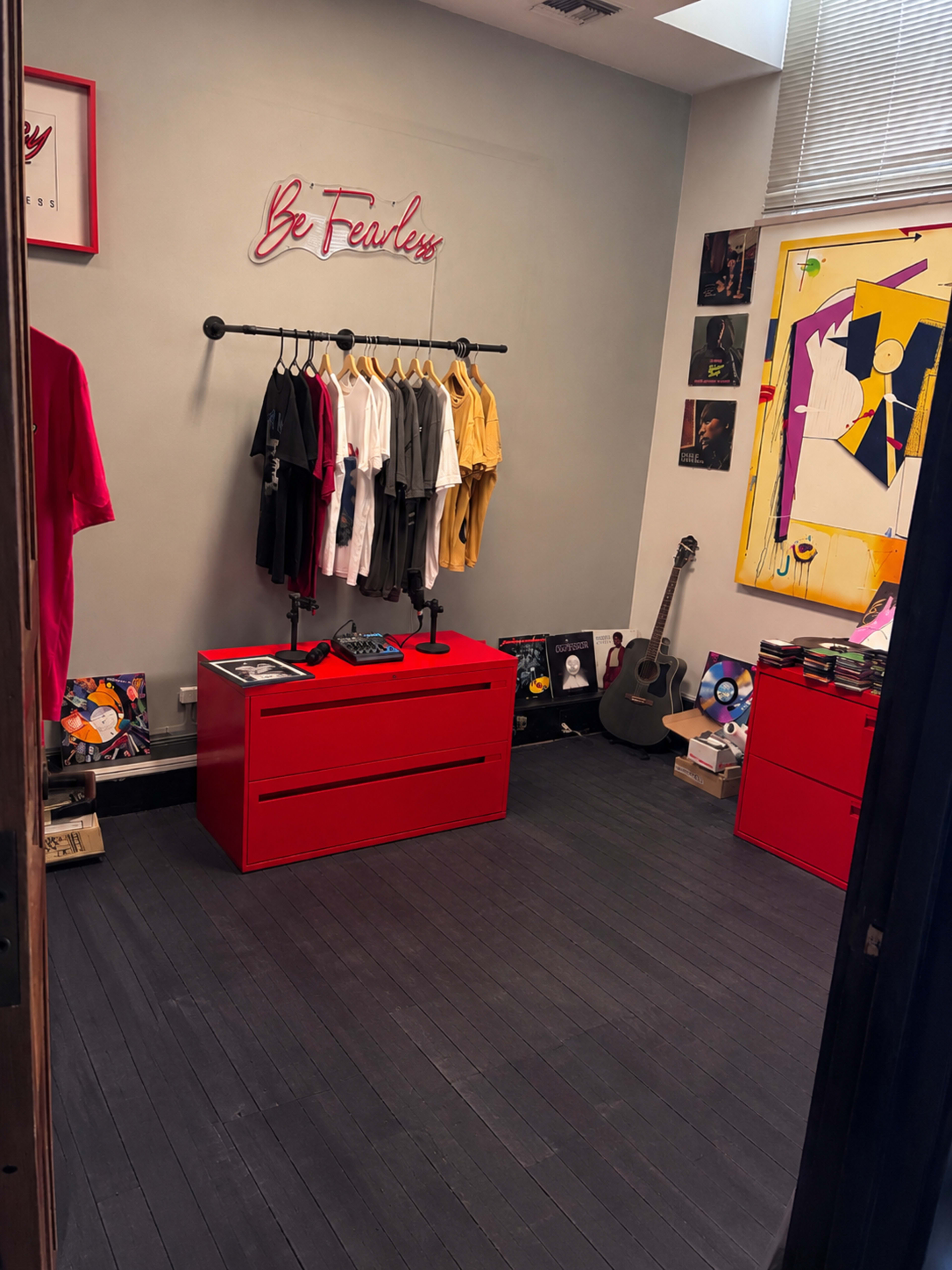 The room features a display of clothing on hangers, colorful art on the walls, a red dresser, and a guitar leaning against the wall.
