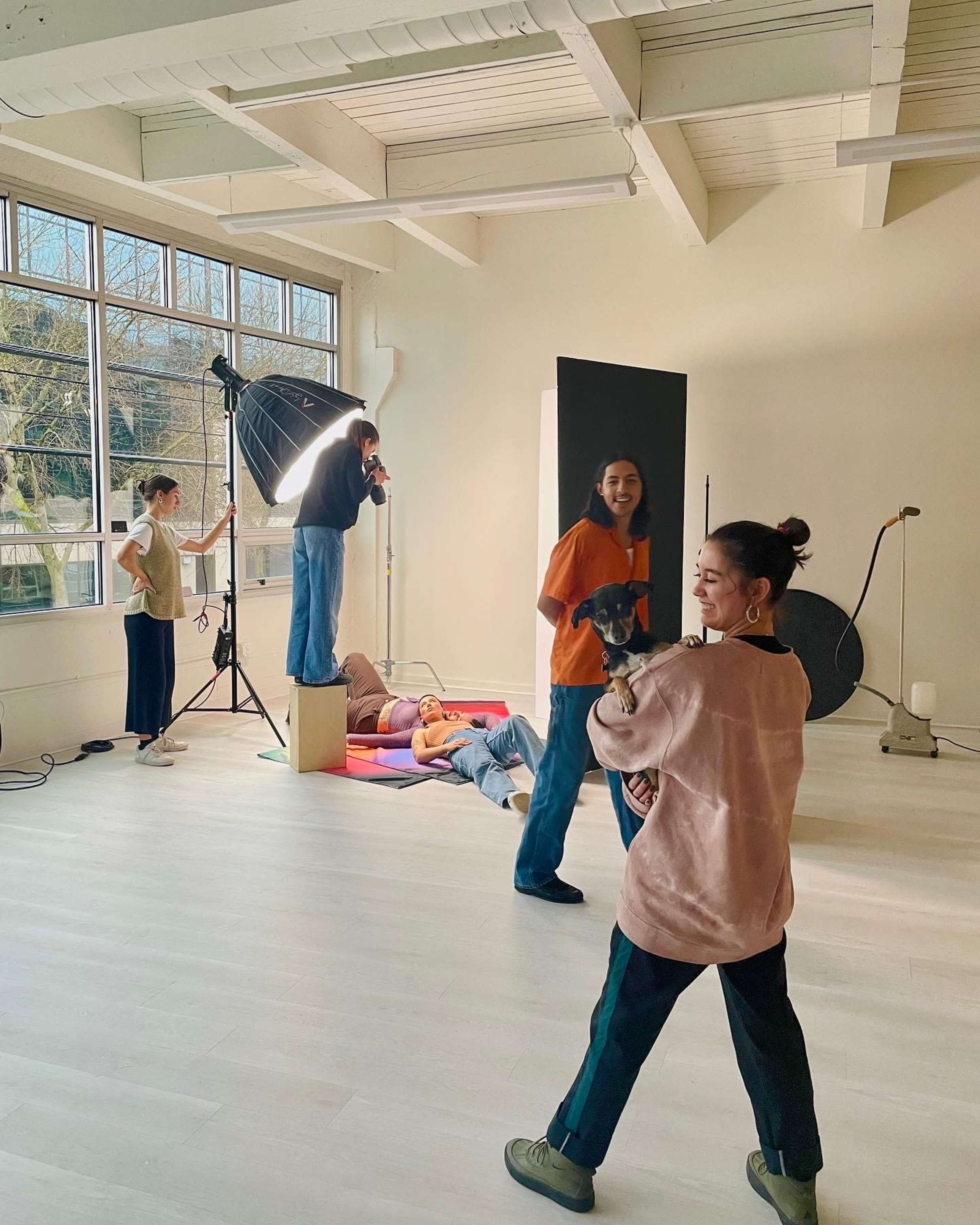 A group of people is engaged in a photoshoot in a well-lit studio, with one person posing on a box while others adjust lighting and equipment.