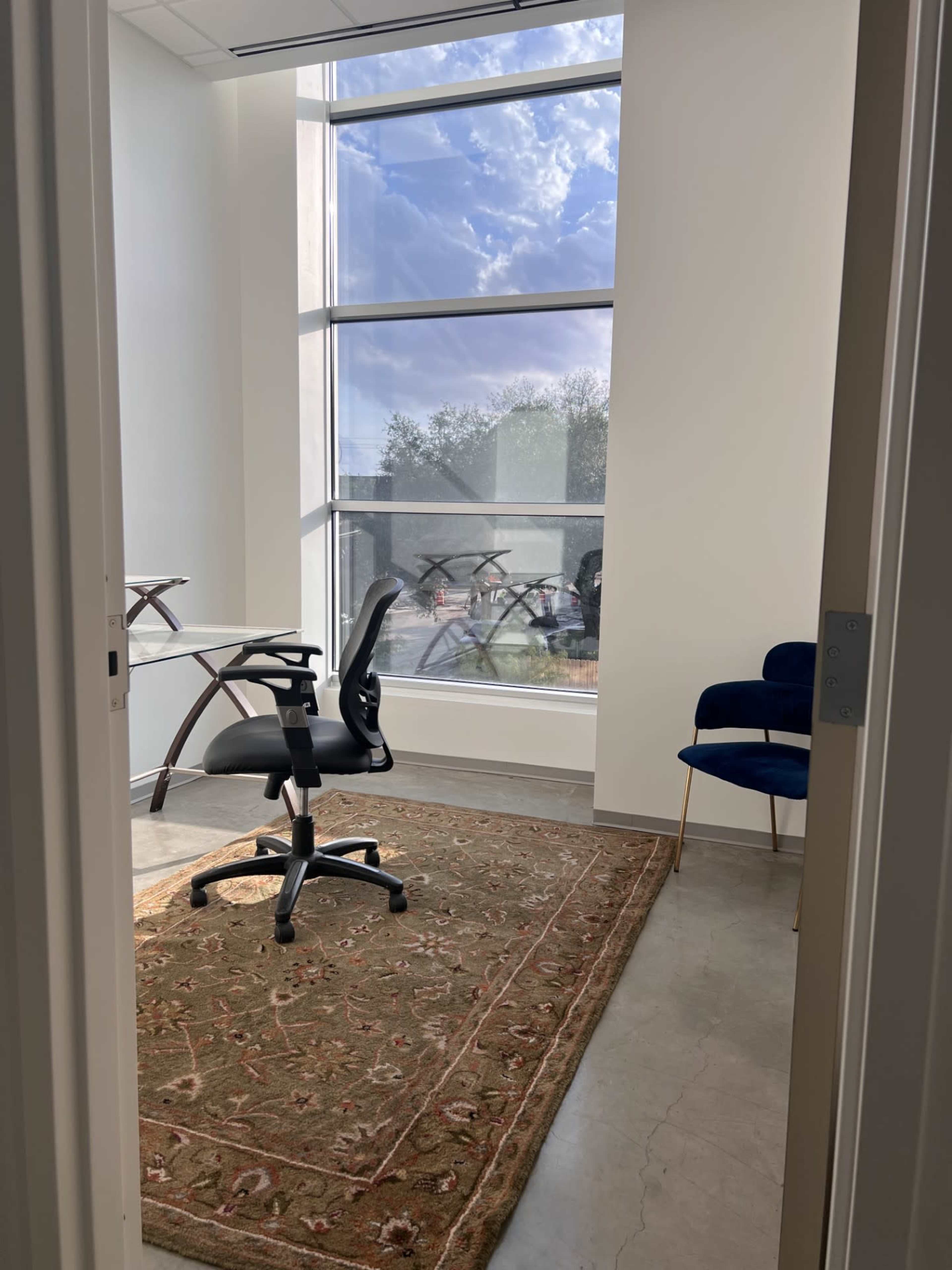 A small office features a desk and a swivel chair on a patterned rug, with large windows allowing natural light to enter.