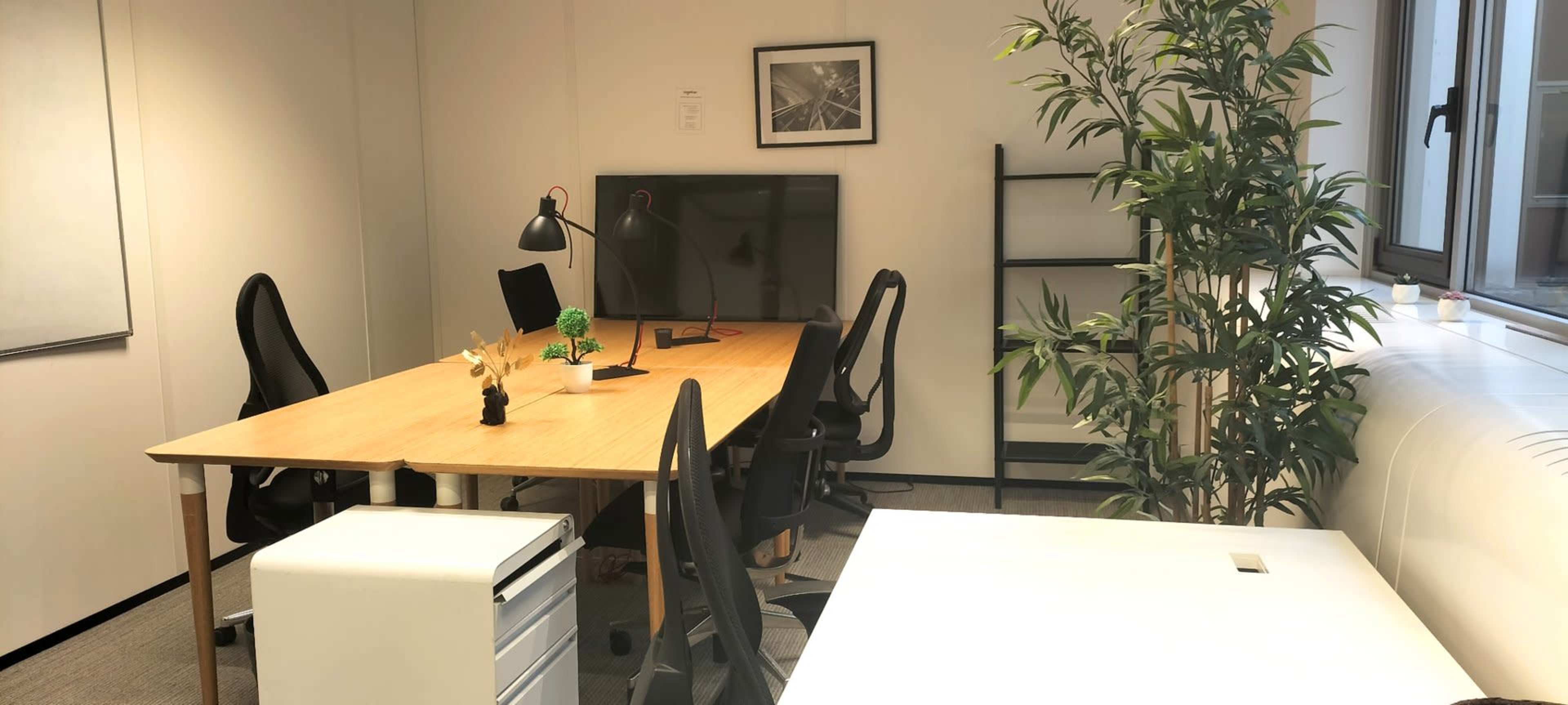 The image shows a modern office space featuring a large wooden table, several black office chairs, a TV monitor, a plant, and a white desk in the corner.