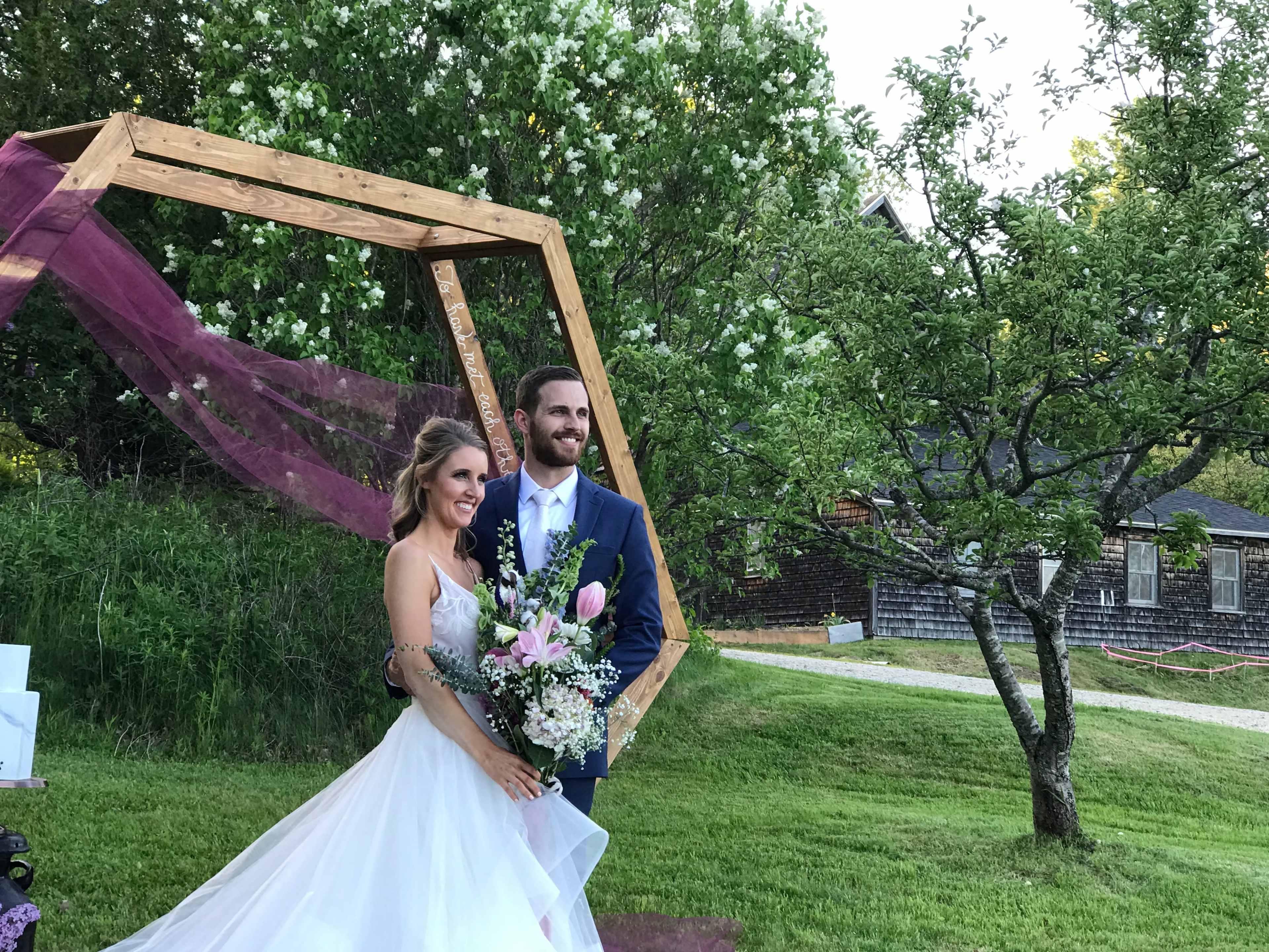 A bride and groom stand together under a wooden arch adorned with fabric and flowers in a grassy outdoor setting.