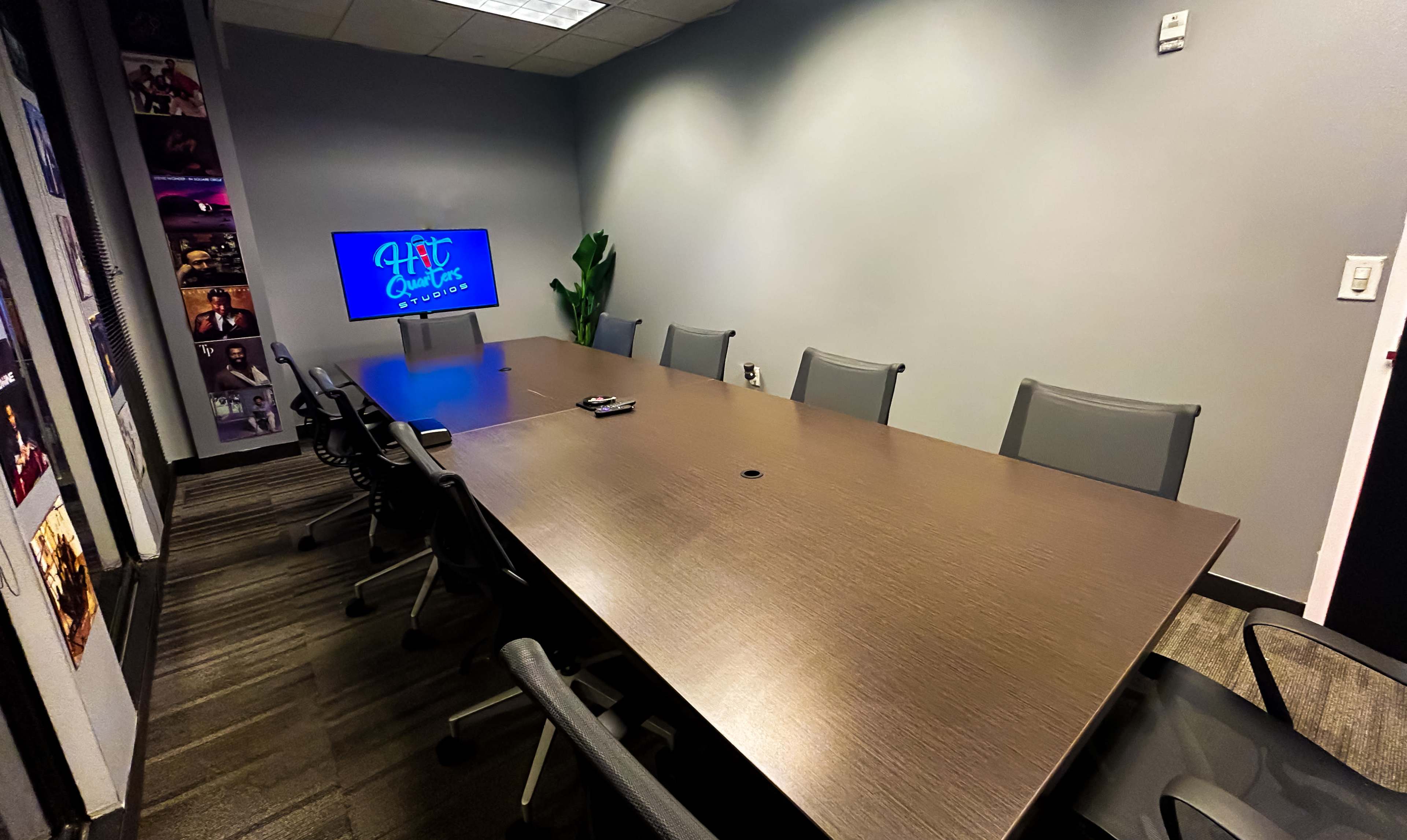 A meeting room with a long wooden table, several chairs, a television display, and a potted plant.