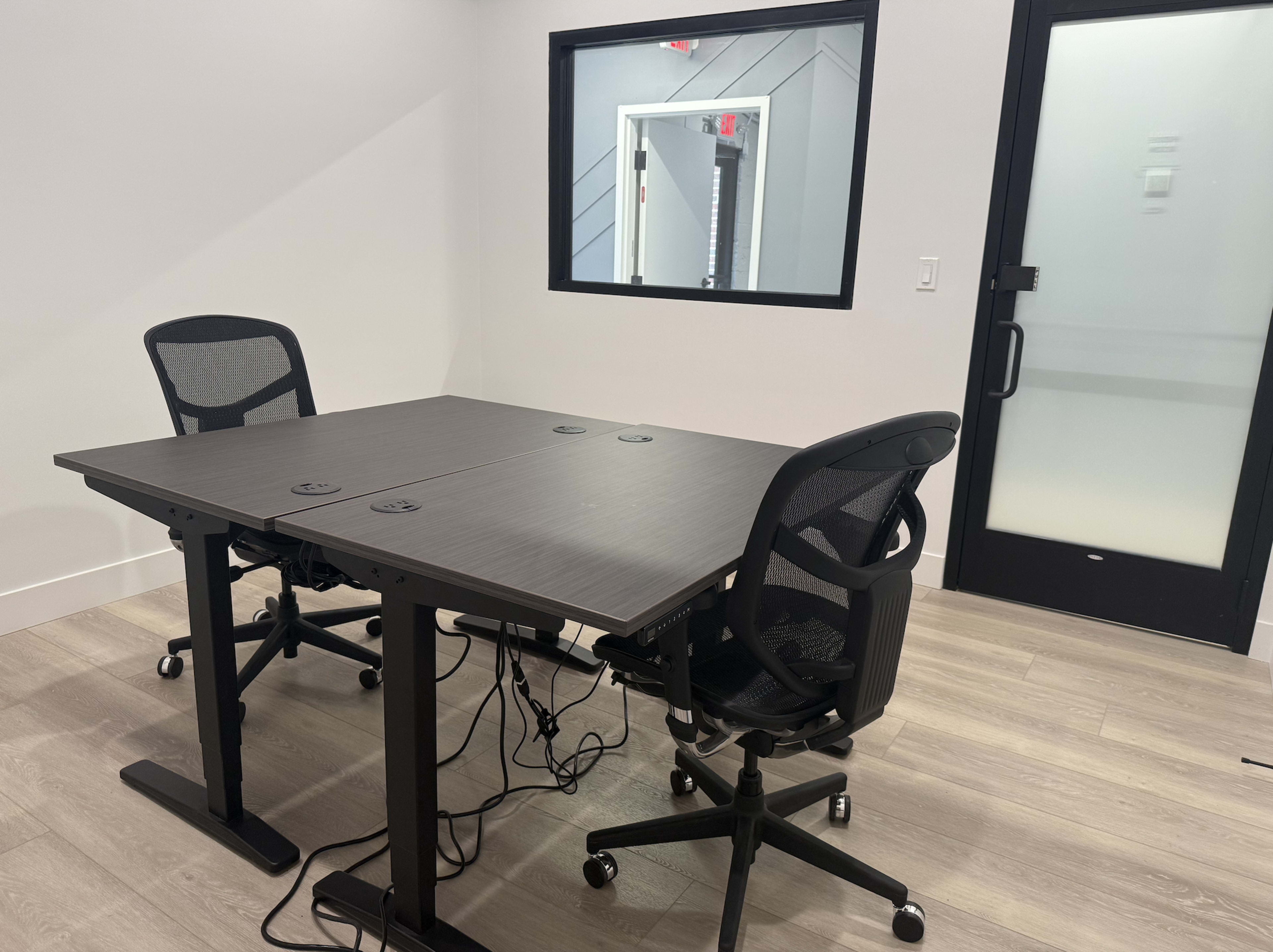 The image shows a small, modern meeting room with a table and two rolling chairs, equipped with a window and a glass door.