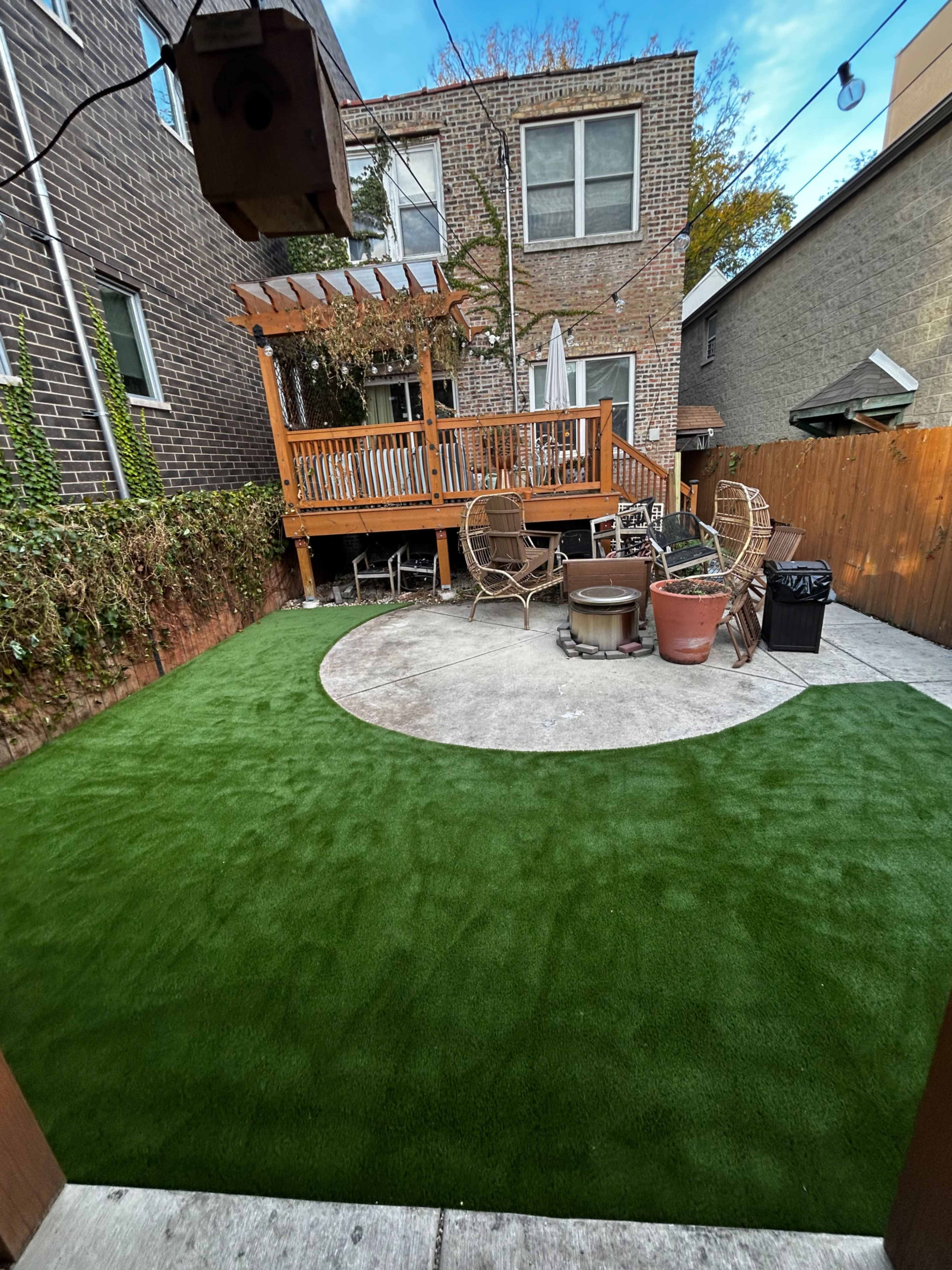 The image shows a backyard featuring a wooden deck, circular patio area with potted plants, and green artificial turf.