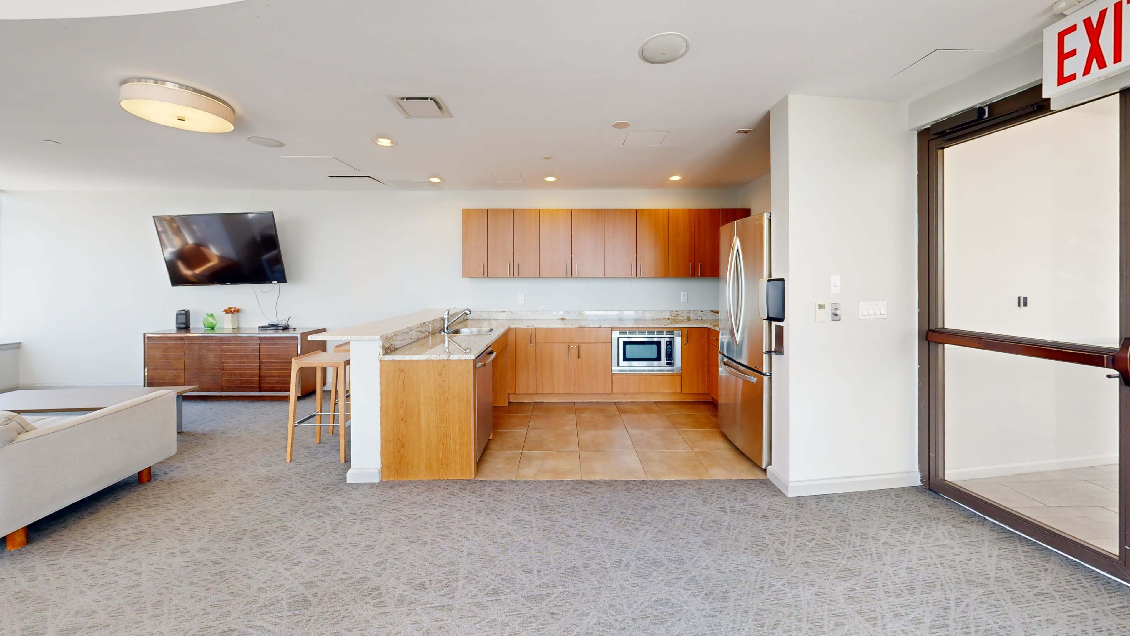The image shows a modern kitchen area with wood cabinets, stainless steel appliances, and a living space featuring a television and exit door.