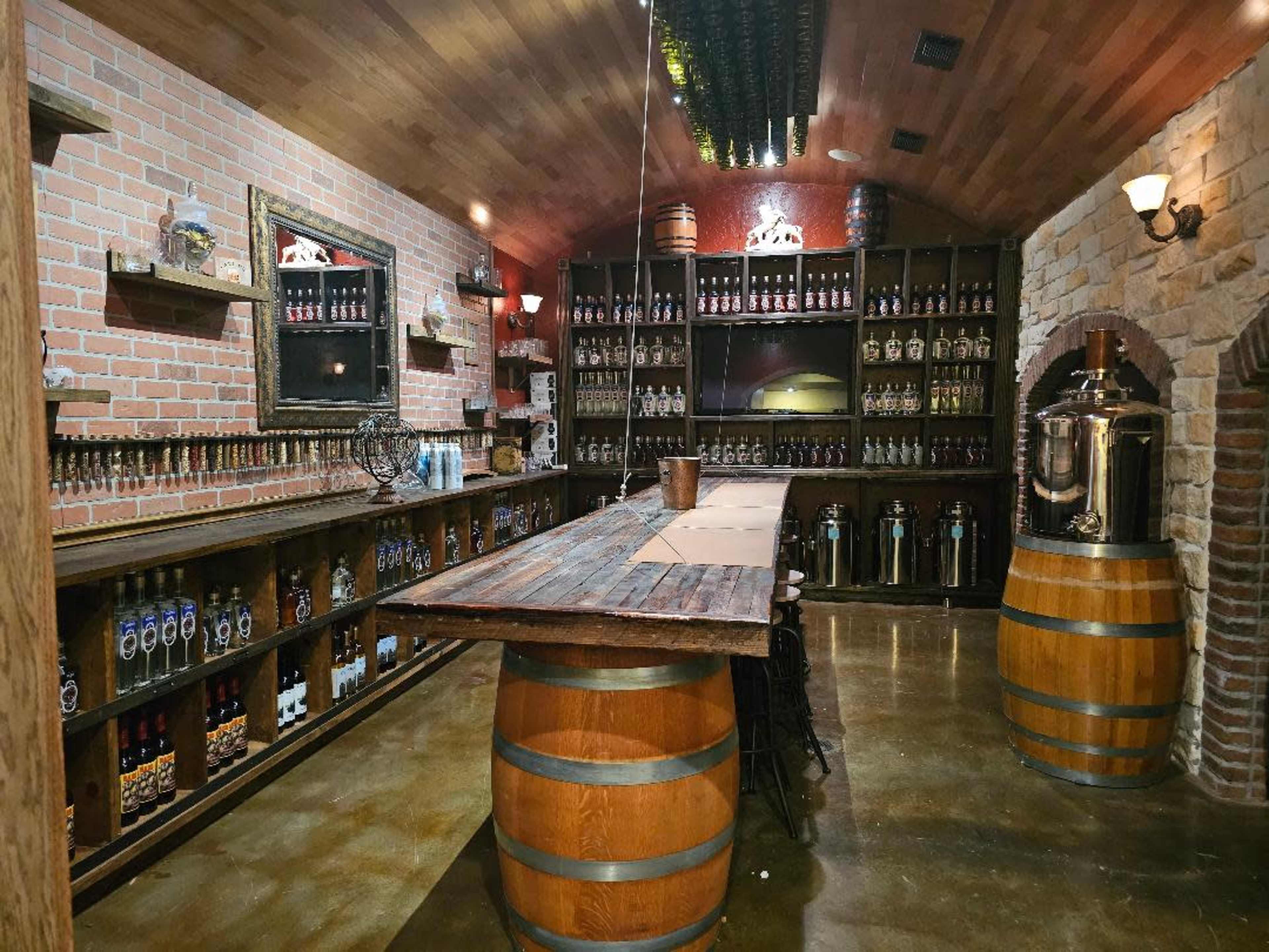 The image shows a dimly lit wine cellar featuring wooden barrels, a long wooden bar, and shelves lined with bottles.