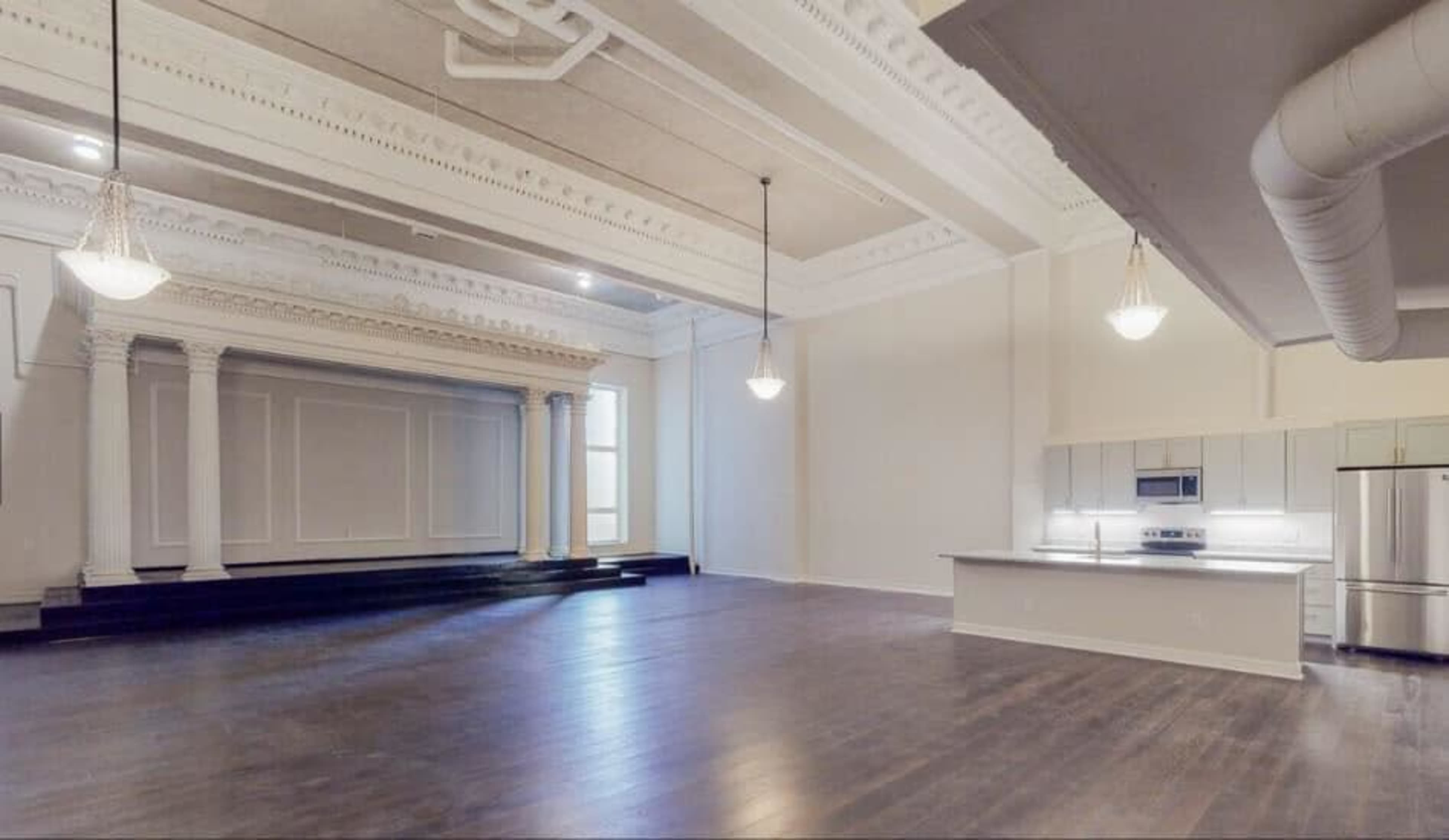 A spacious room with a high ceiling, featuring a decorative columned stage area and an open kitchen area with modern appliances.