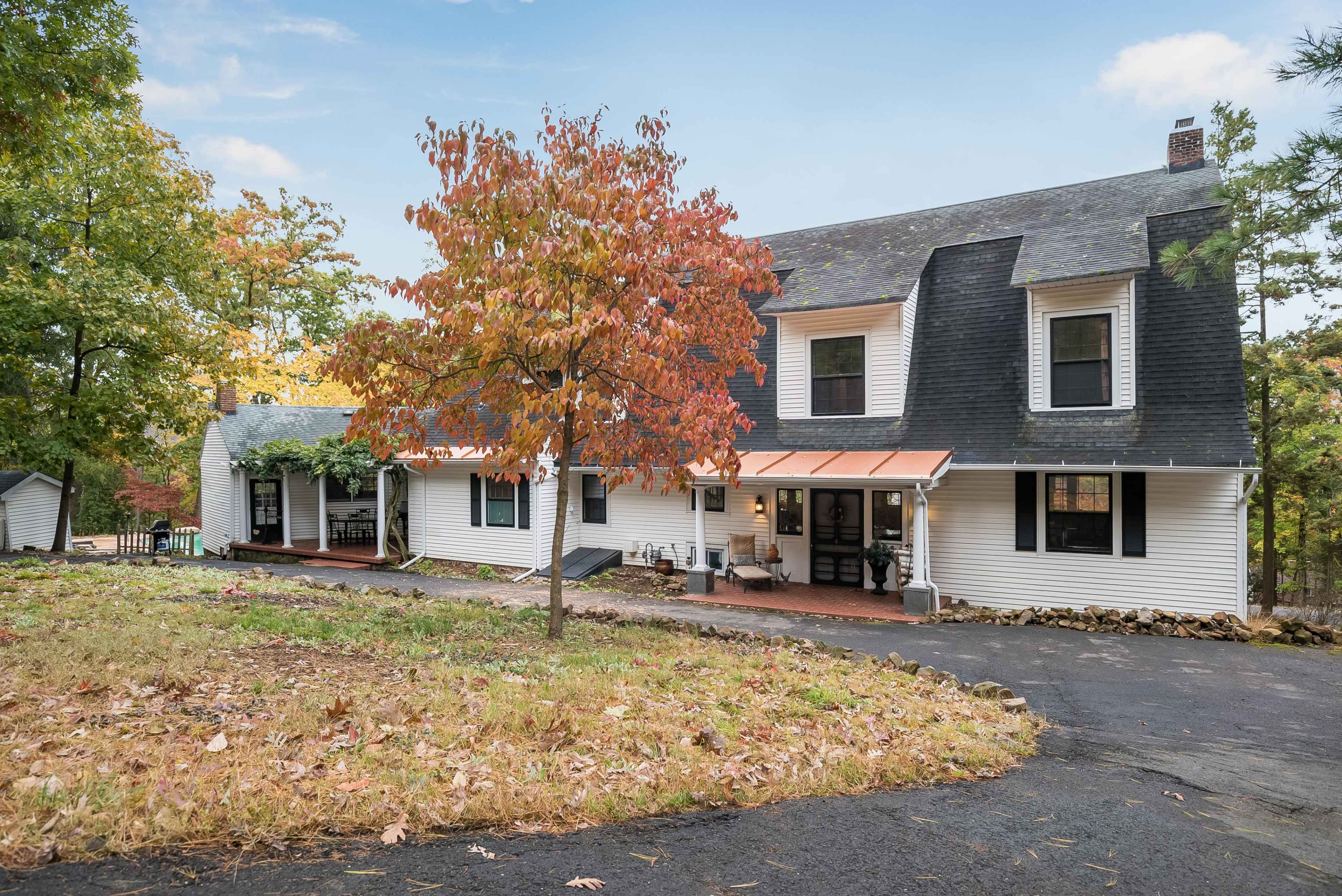 A large white house with a black roof, set in a wooded area with autumn foliage and a gravel driveway.