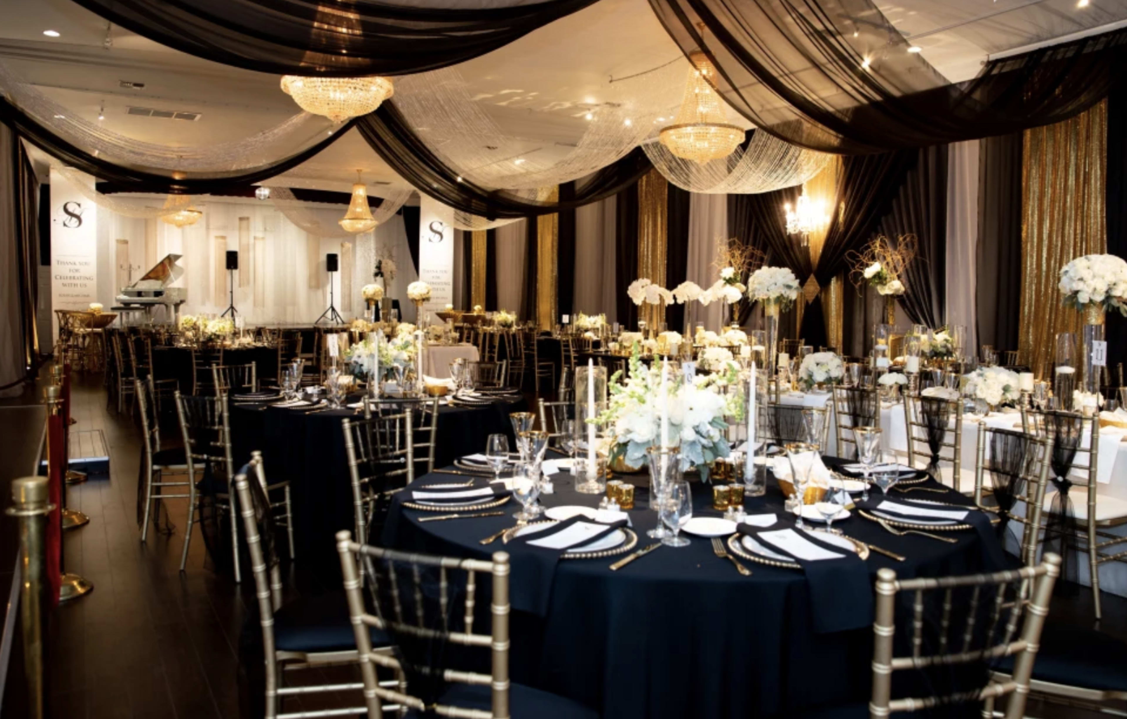 A elegantly decorated banquet hall with round tables set for a formal event, featuring floral centerpieces and draped fabric overhead.