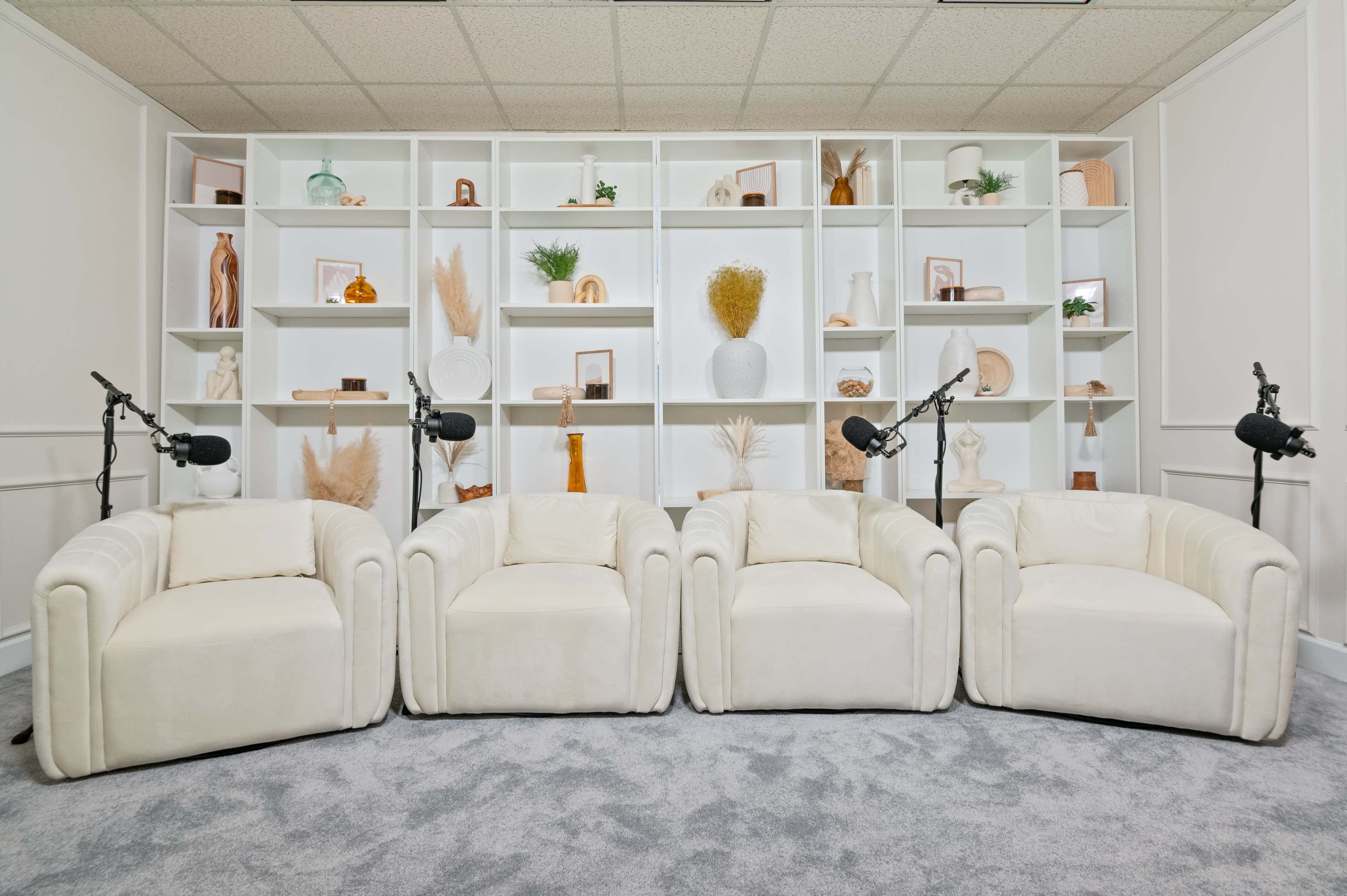 Four white upholstered chairs arranged in front of a decorative wall shelf filled with various items.
