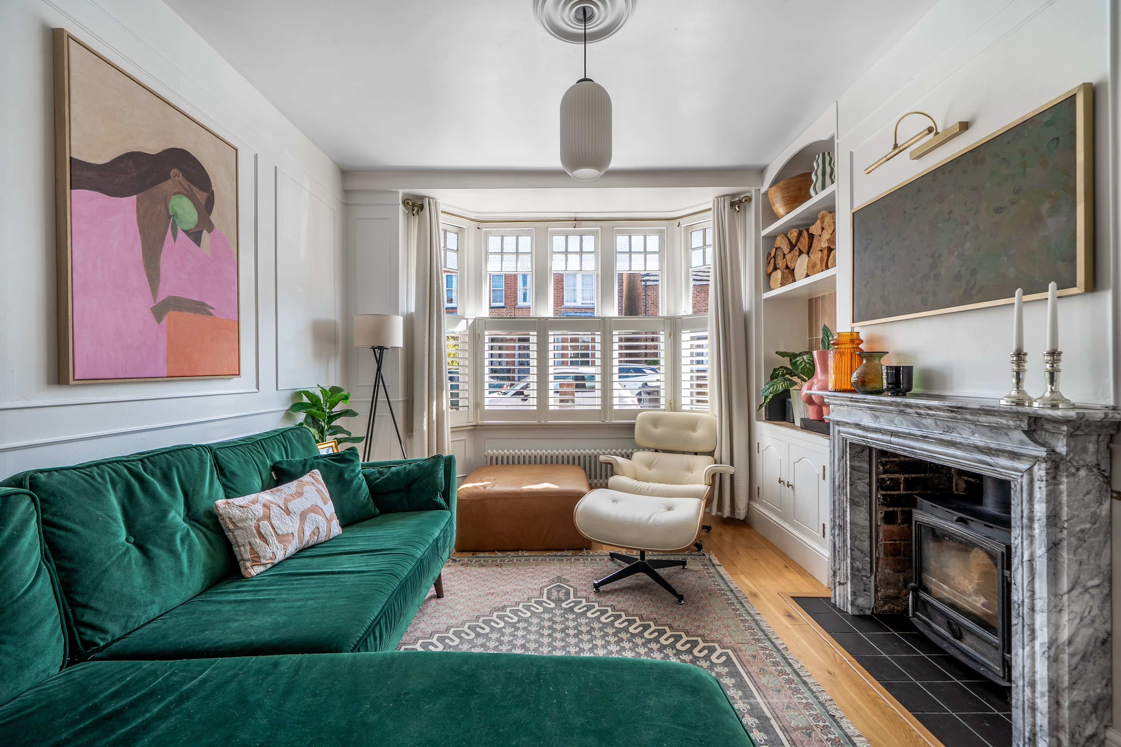 A stylish living room features a green sofa, an armchair, a marble fireplace, a large window with shutters, and decorative artwork on the walls.