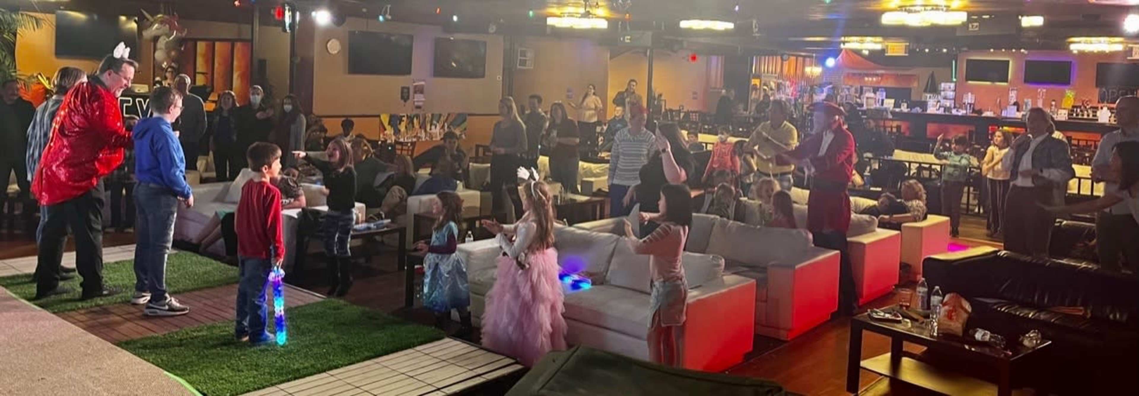 A lively event is taking place in a spacious venue filled with guests of various ages, some seated at tables while others participate in activities on a stage area with colorful lighting.