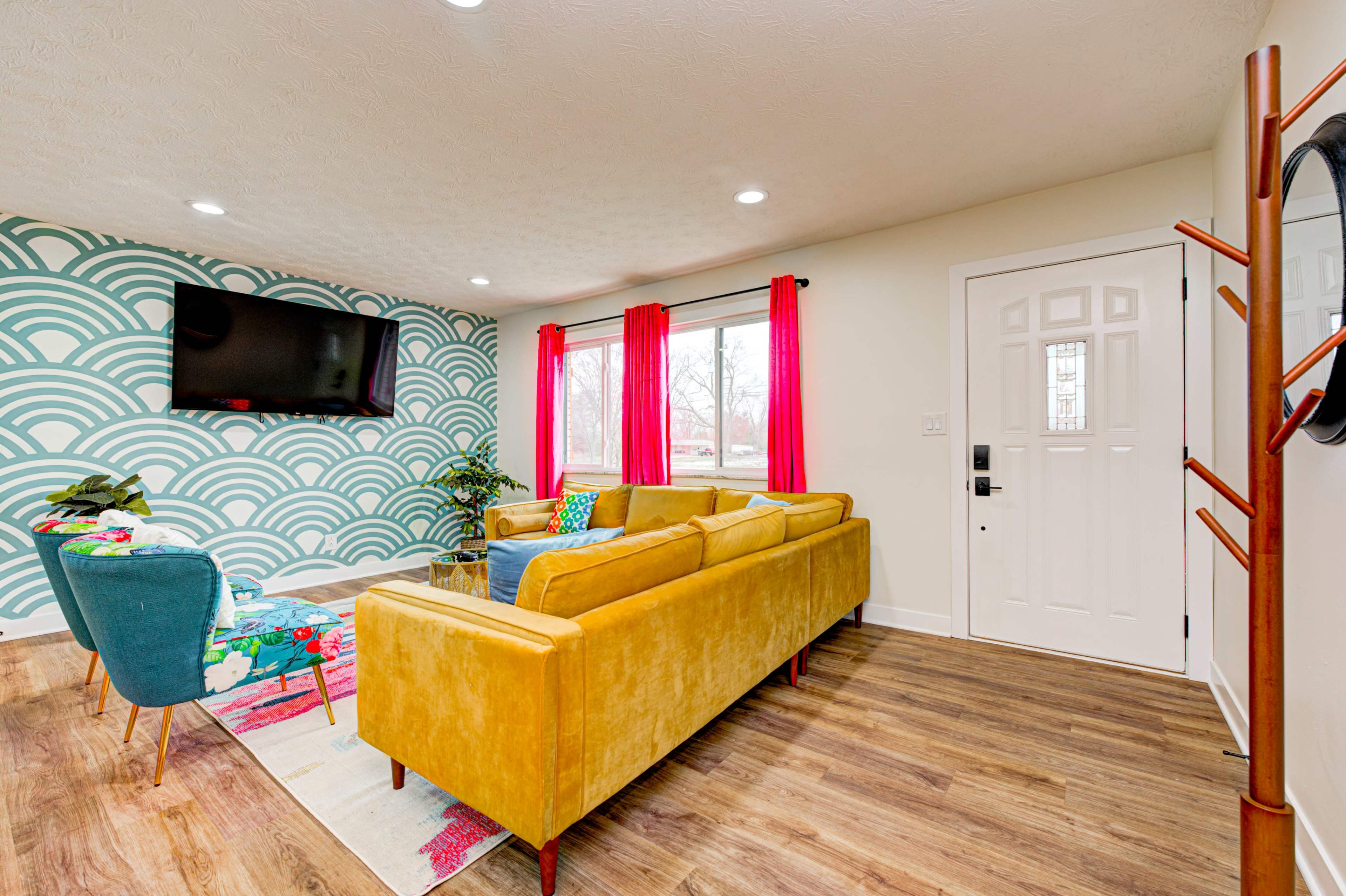 The living room features a yellow couch, a green chair, a wall with a teal wave pattern, and large windows with red curtains.