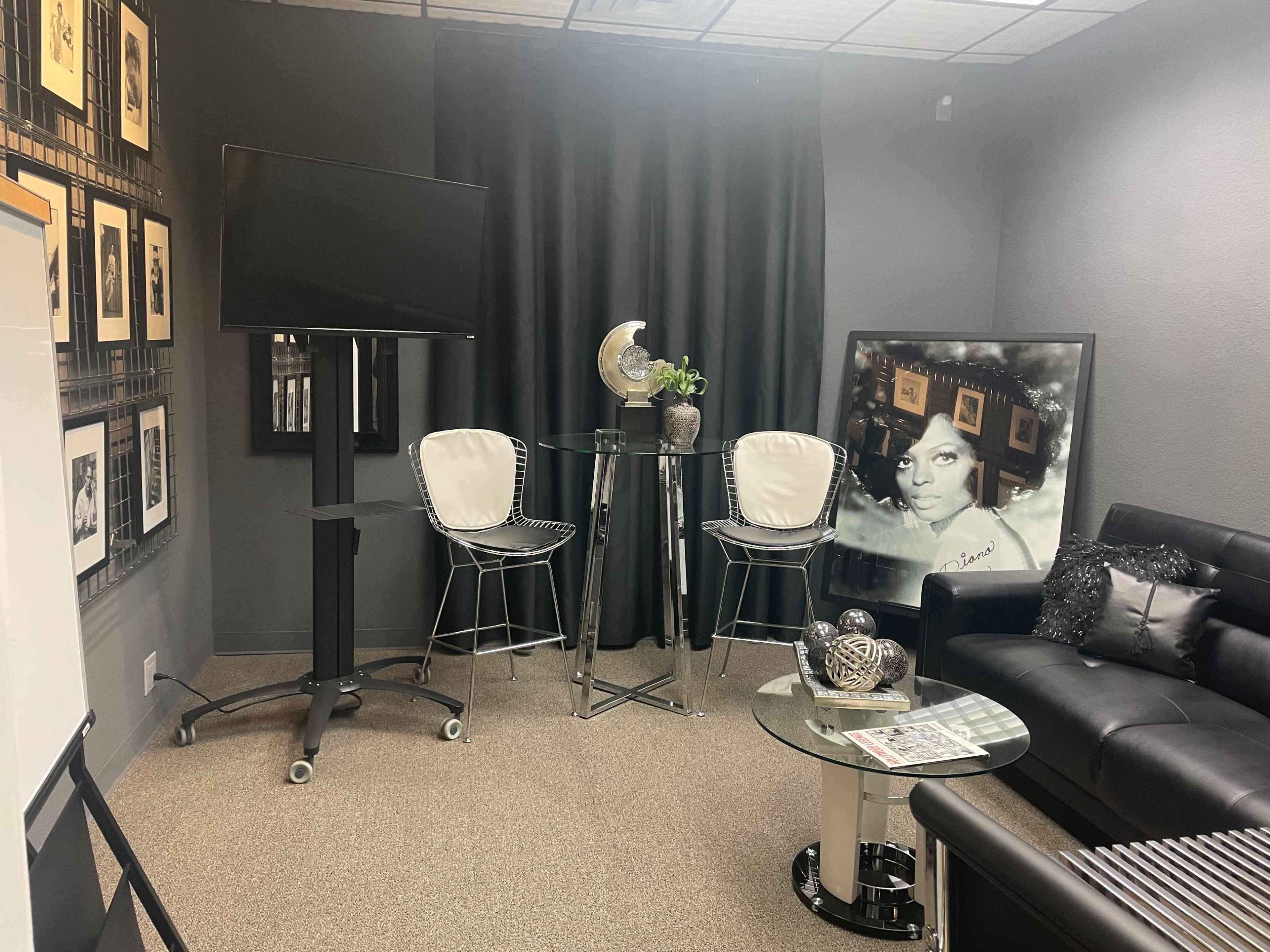 The image shows a small, modern lounge area featuring a black sofa, a glass table with high stools, a mounted television, and framed pictures on the walls.