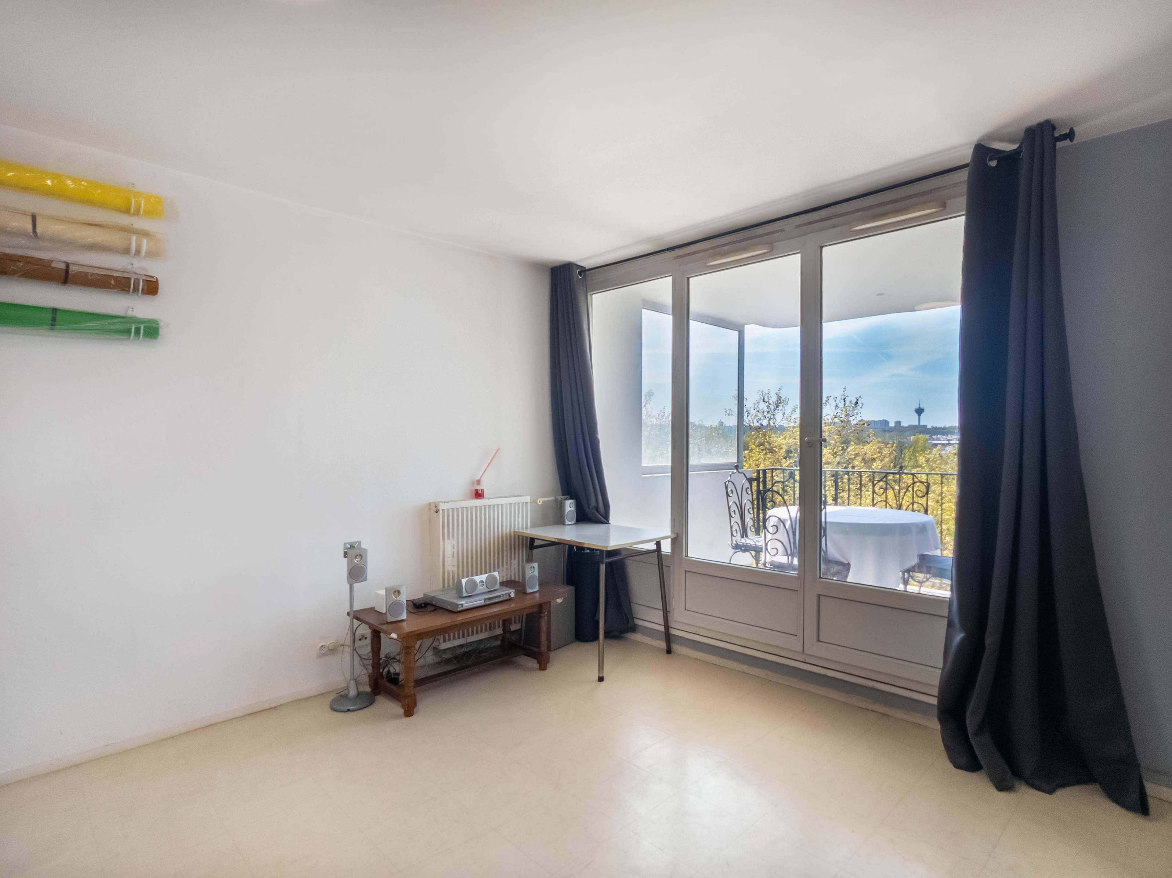 The room features large windows with a view of a balcony and is furnished with a table, a chair, and a radiator.