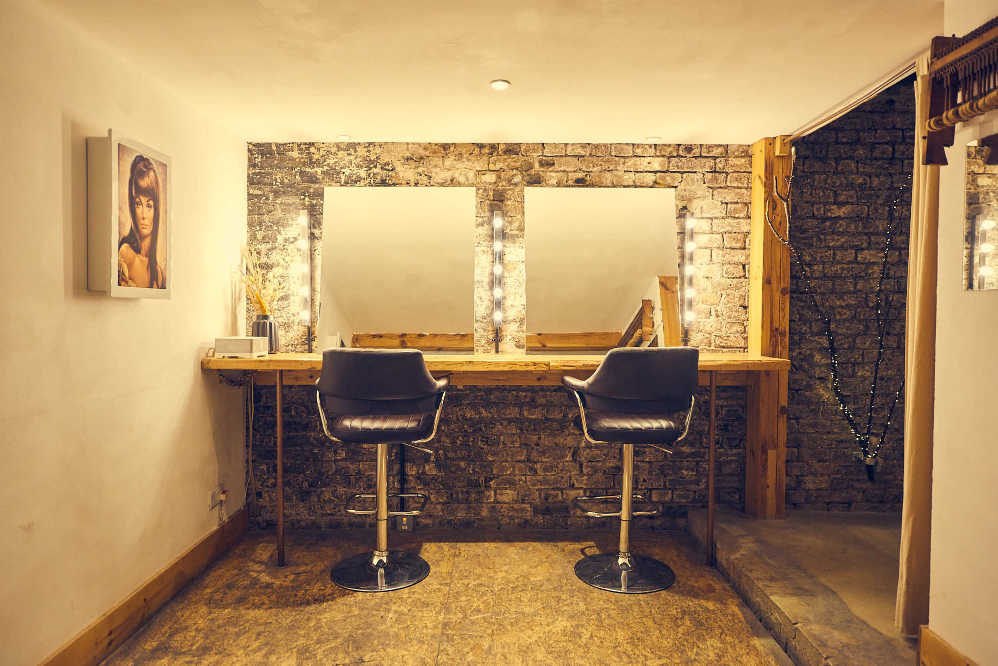 A makeup station with two black bar stools, a wooden countertop, and illuminated mirrors against a brick wall.