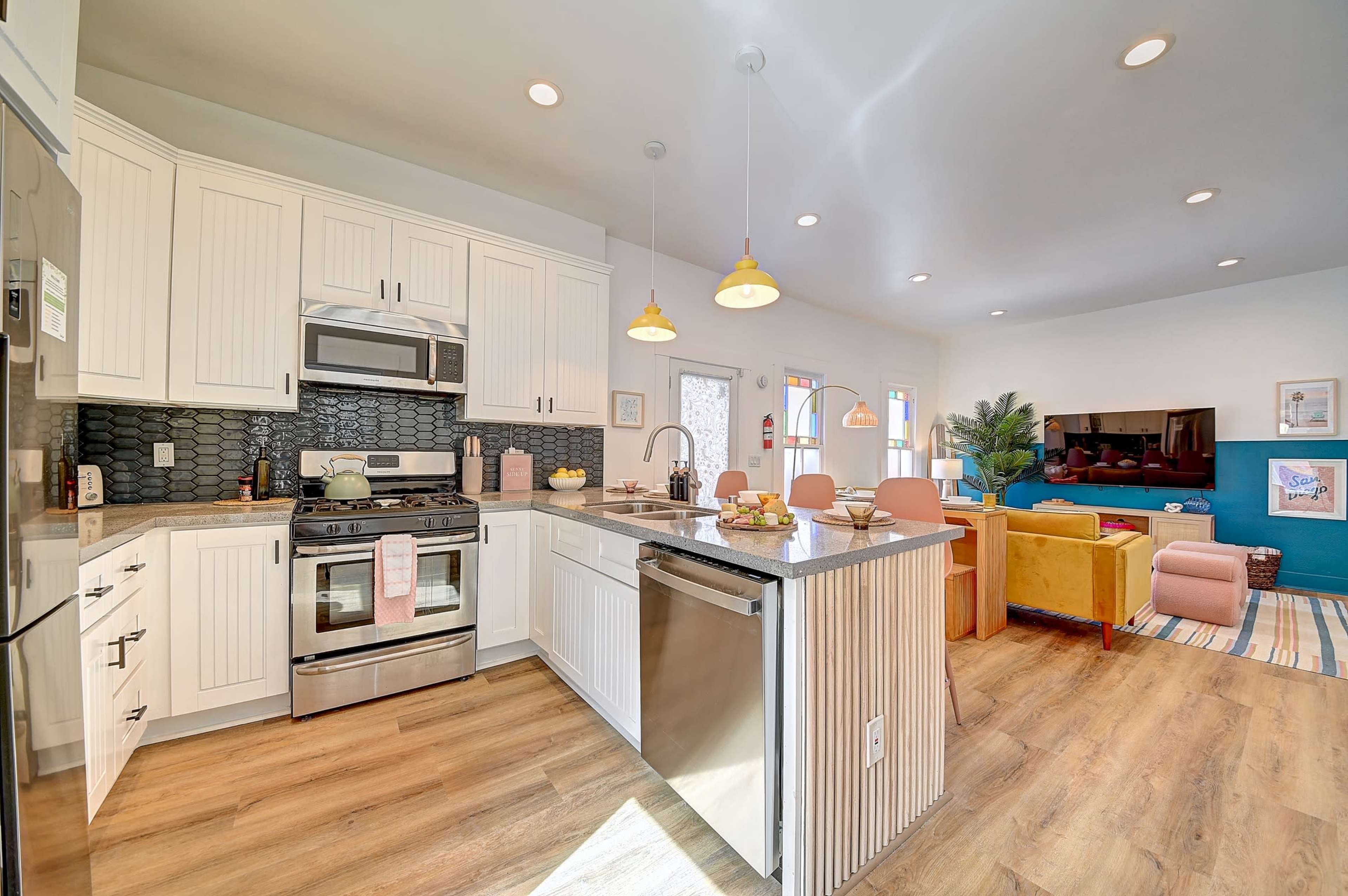 The image shows a modern kitchen with white cabinetry, stainless steel appliances, and a granite countertop, leading to a living area with colorful furniture and decorative art.