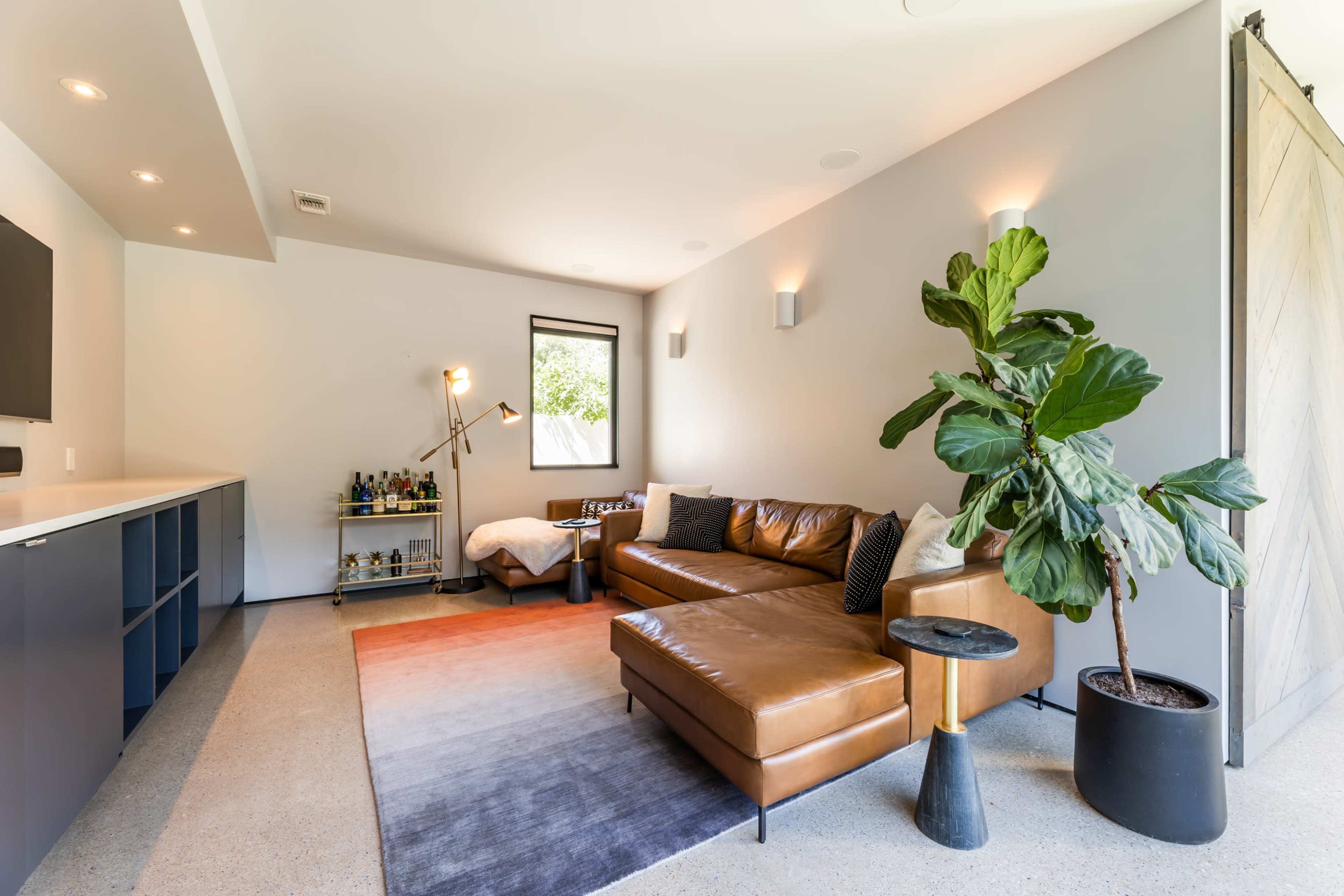A modern living room featuring a brown leather sectional sofa, a round side table, a large potted plant, and a bar area with shelves in the background.