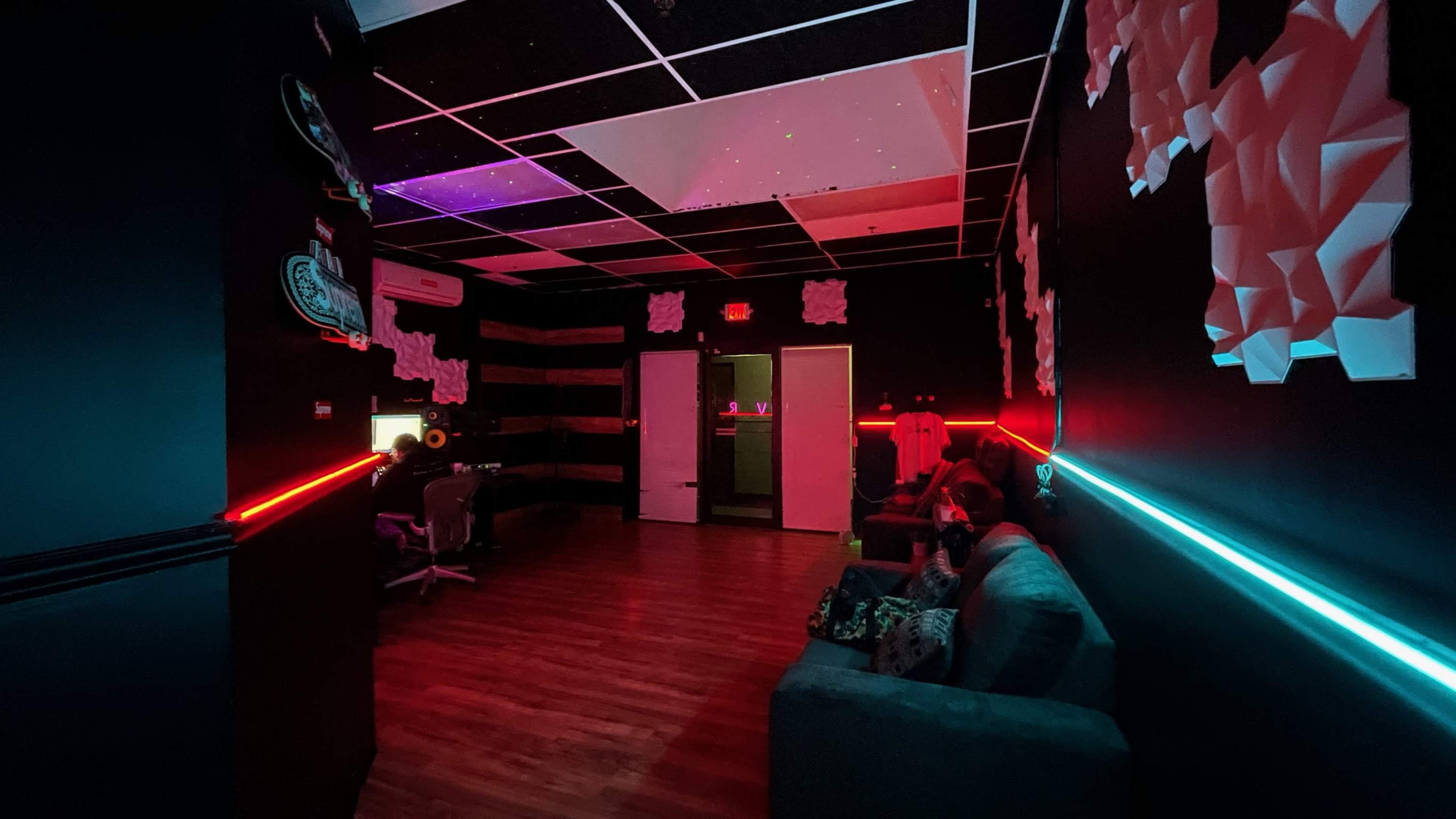 The image shows a dimly lit room with dark walls, colorful LED lights, and modern furniture, including seating and a workspace.