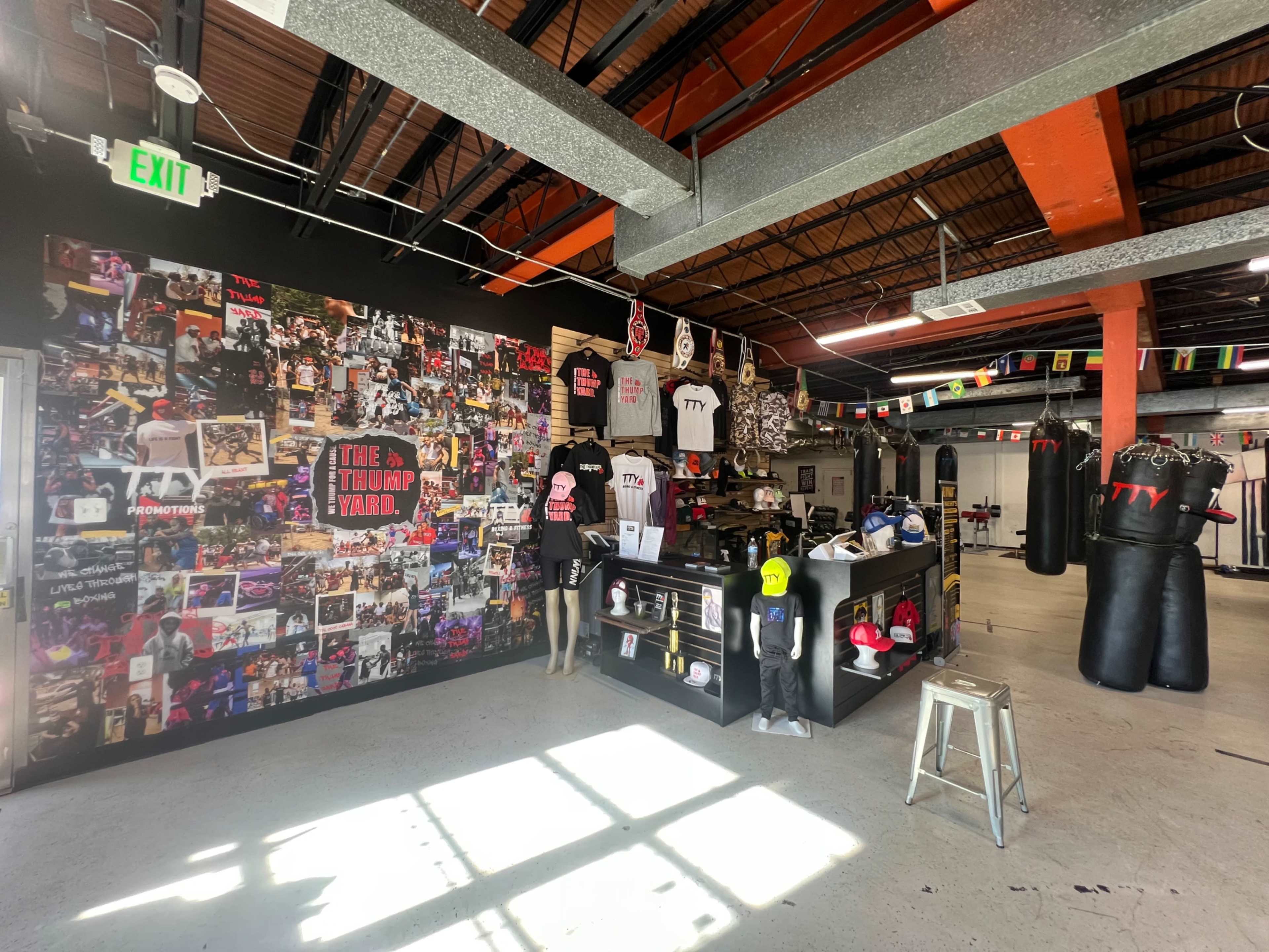 The image shows a boxing gym interior featuring a merchandise display area, a wall covered with collages of photographs, and a variety of boxing gear and apparel hanging from the ceiling.