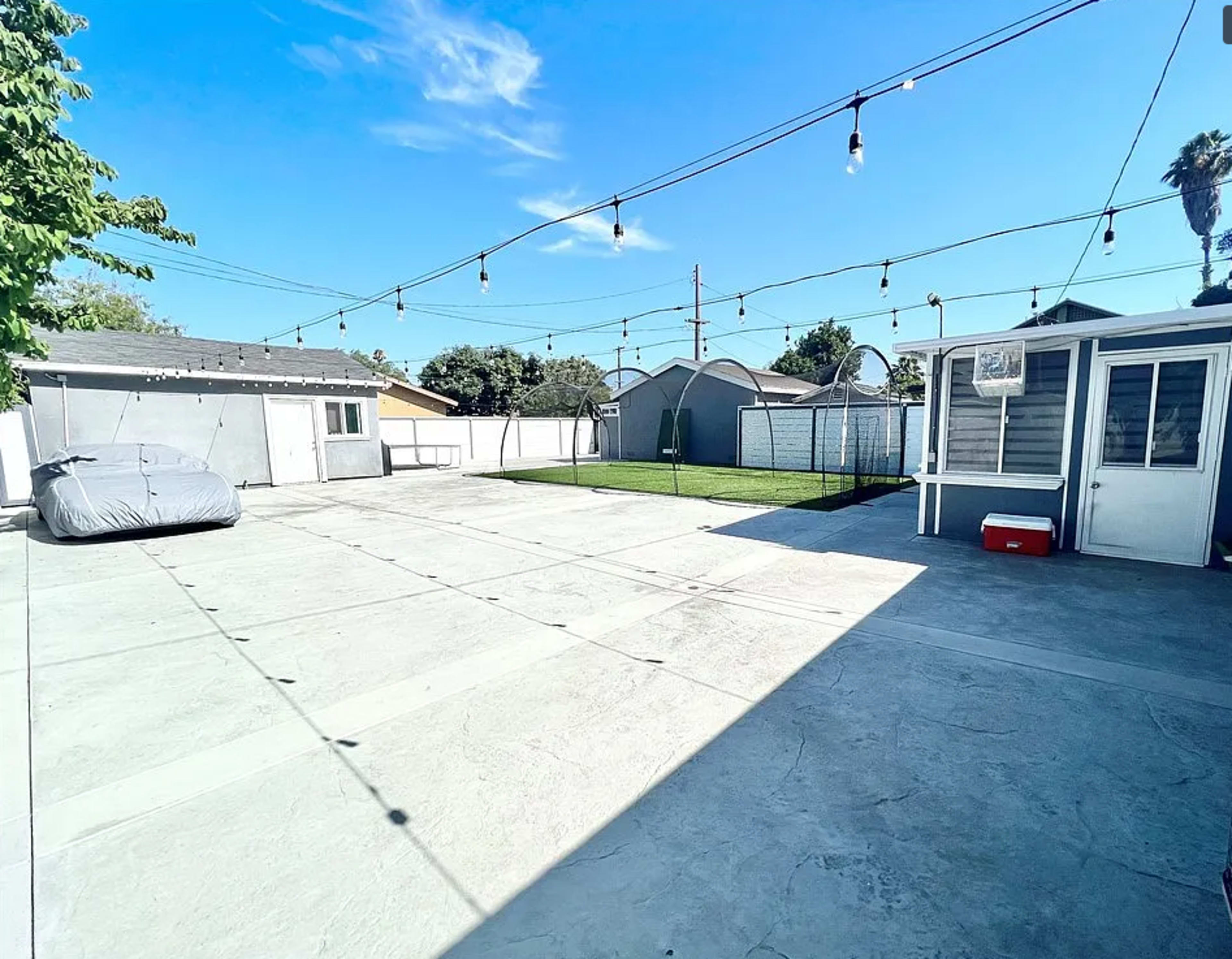 The image shows a spacious outdoor area with a concrete surface, string lights overhead, a gray cover over a vehicle, a green lawn in the background, and a small structure on the right side.