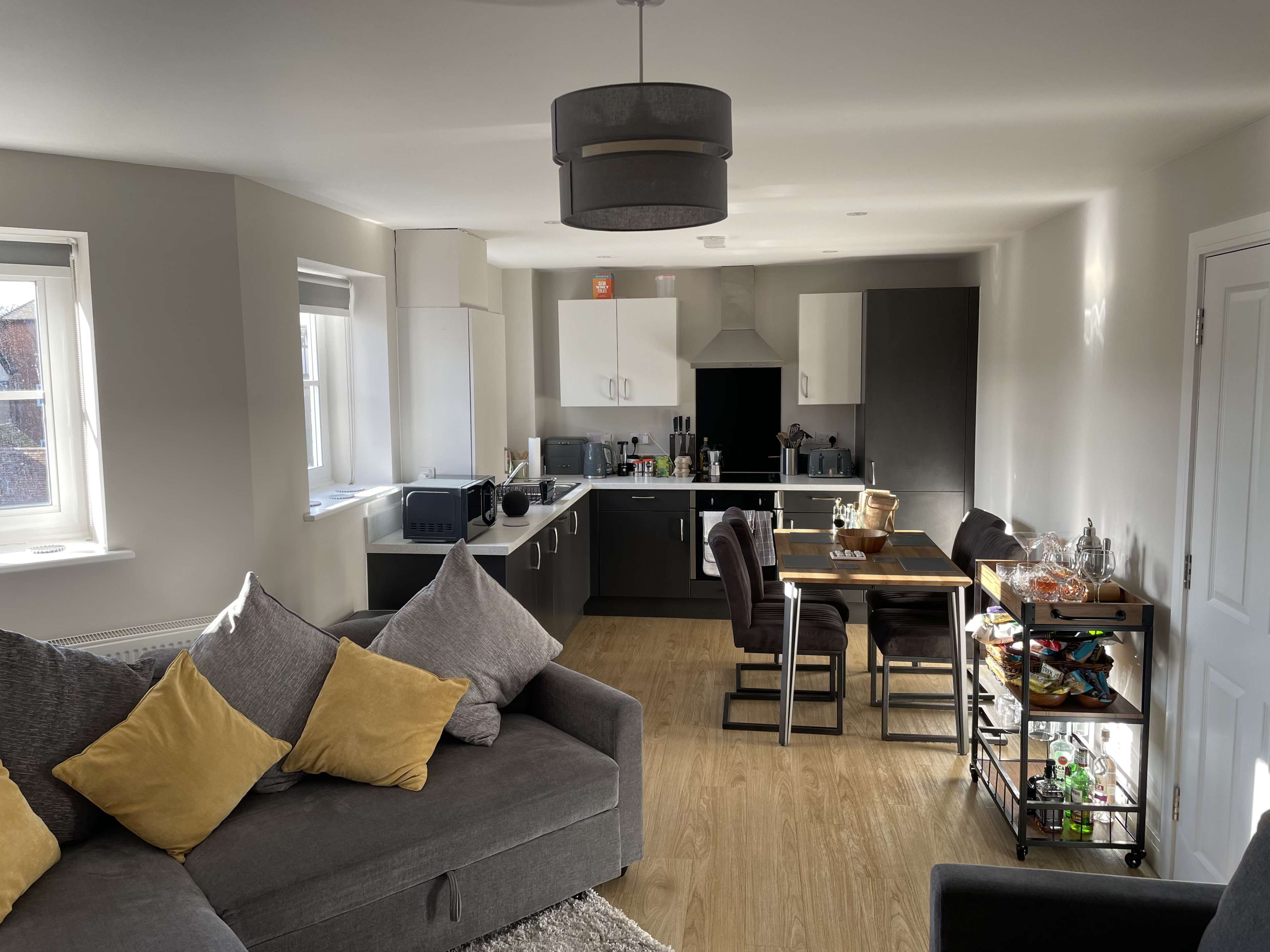 The image shows a modern open-plan living space featuring a gray sectional sofa, a kitchen with dark cabinetry, and a dining area with a wooden table and chairs.