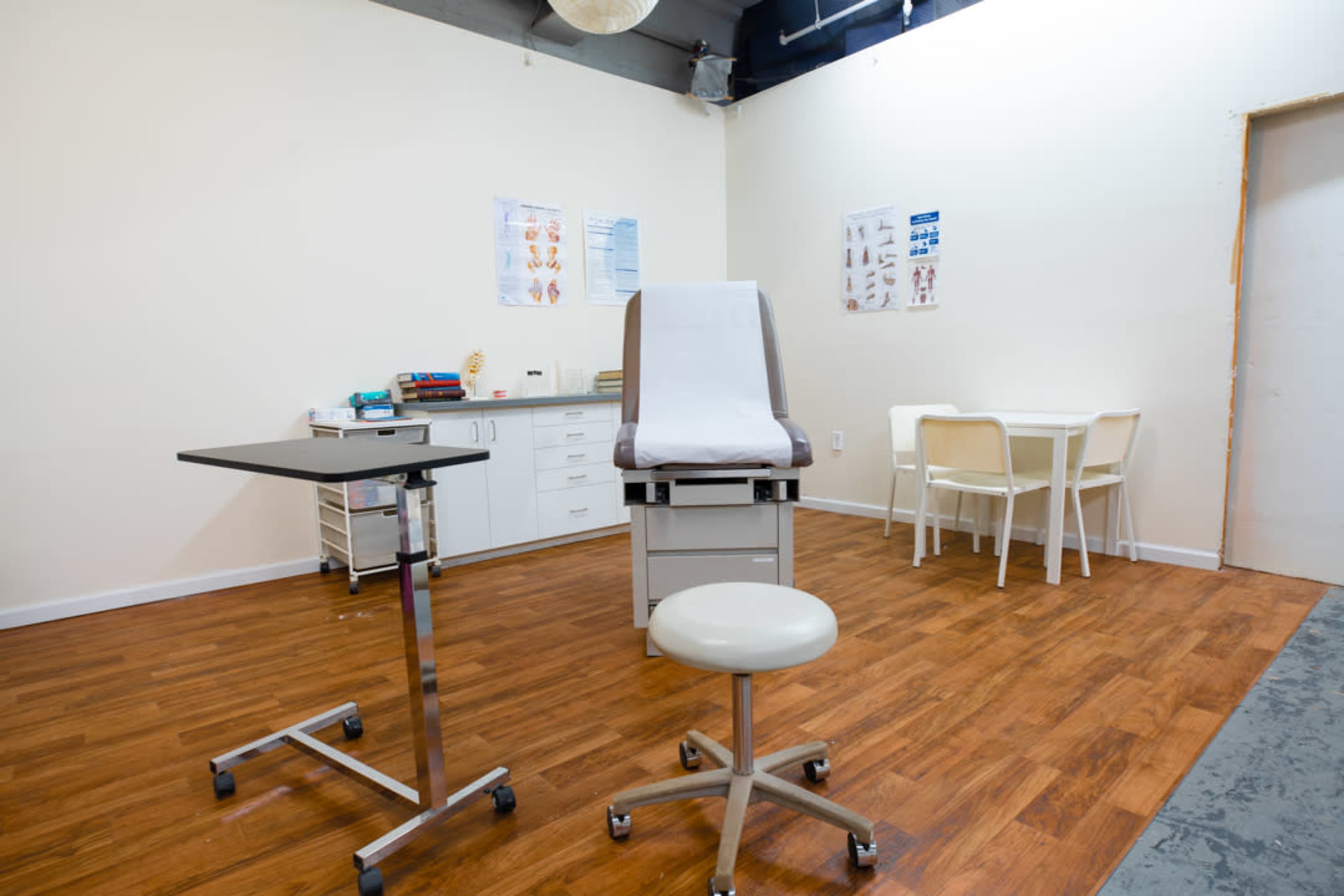 The image shows a small medical examination room with a treatment table, a rolling stool, a movable table, and a set of chairs.