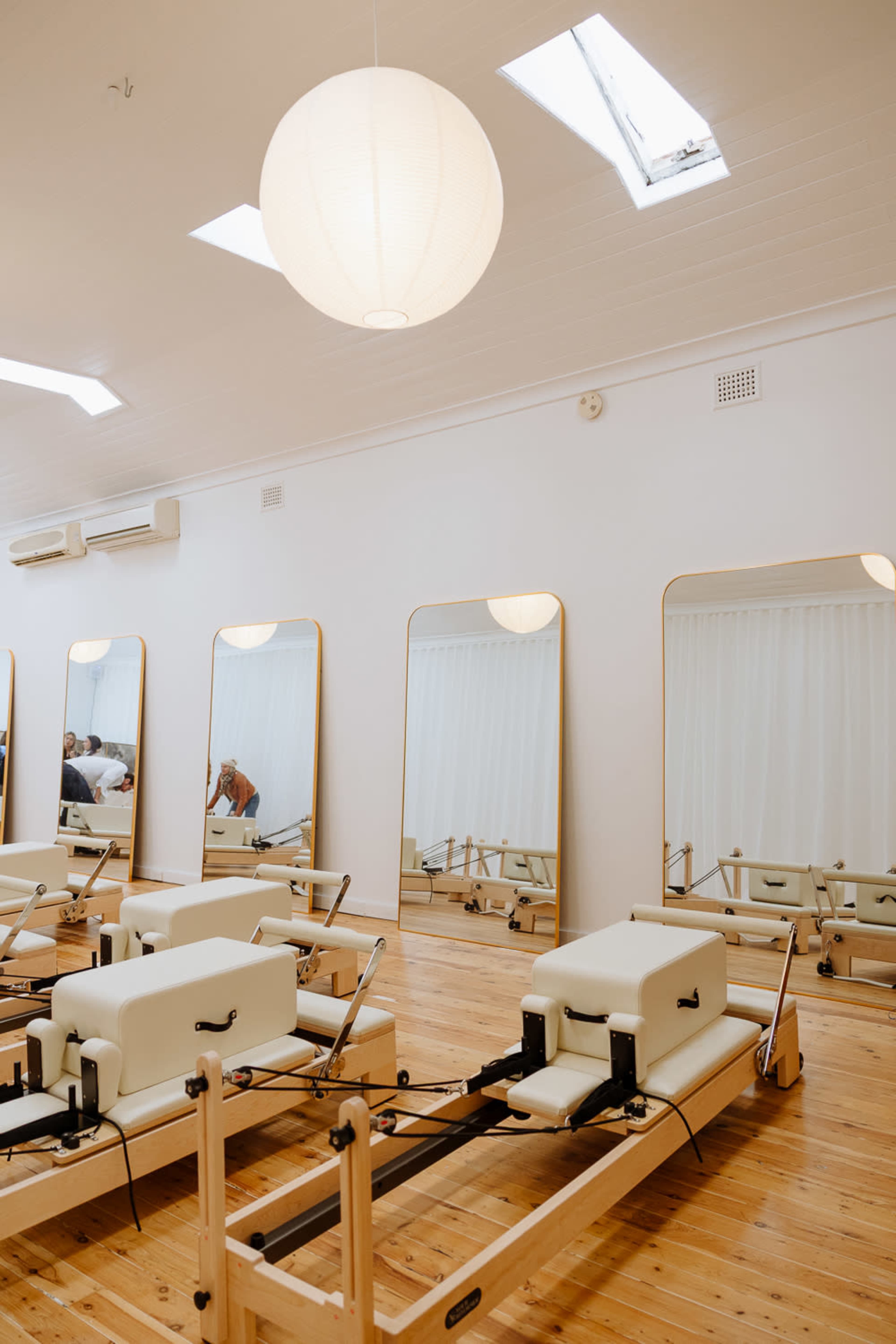 The image shows a well-lit pilates studio featuring several reformer machines arranged on a polished wooden floor, with large mirrors and a circular light fixture above.