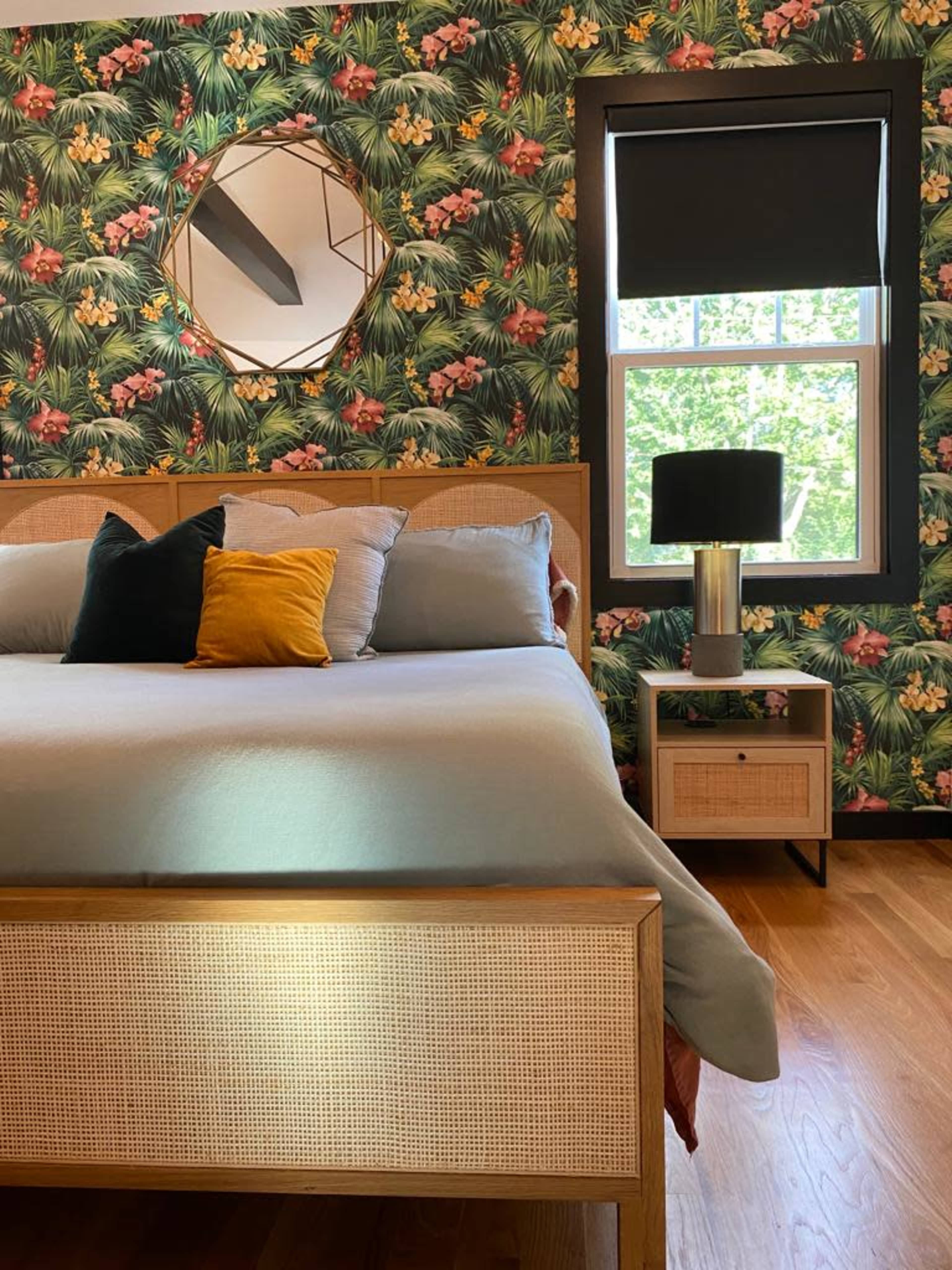 A bedroom featuring a bed with a light blue duvet and colorful pillows, surrounded by floral wallpaper and a bedside table with a lamp.