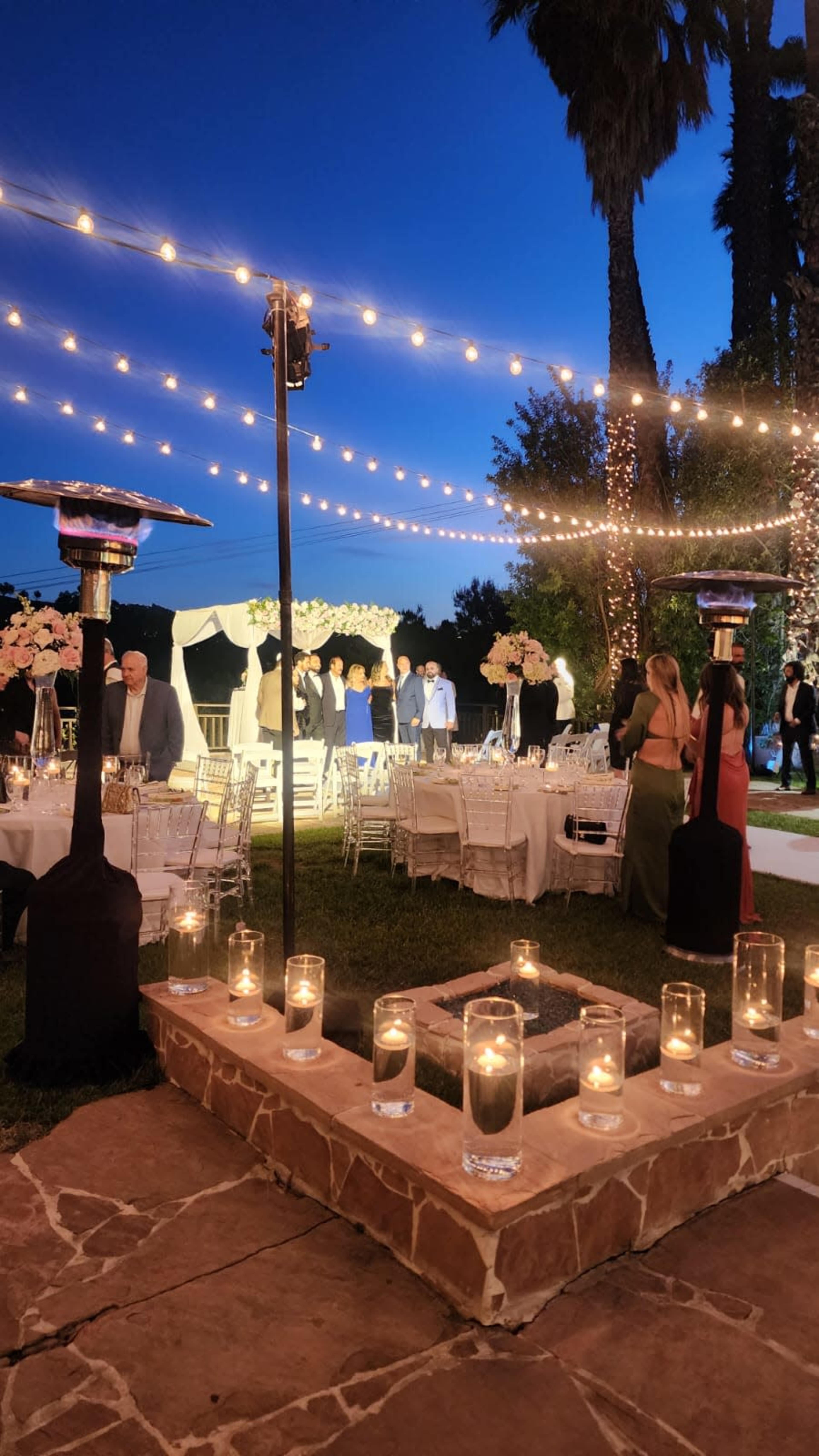 The scene depicts a beautifully arranged outdoor event at night, with tables set for dining, string lights overhead, and guests mingling around a patio area adorned with candles.