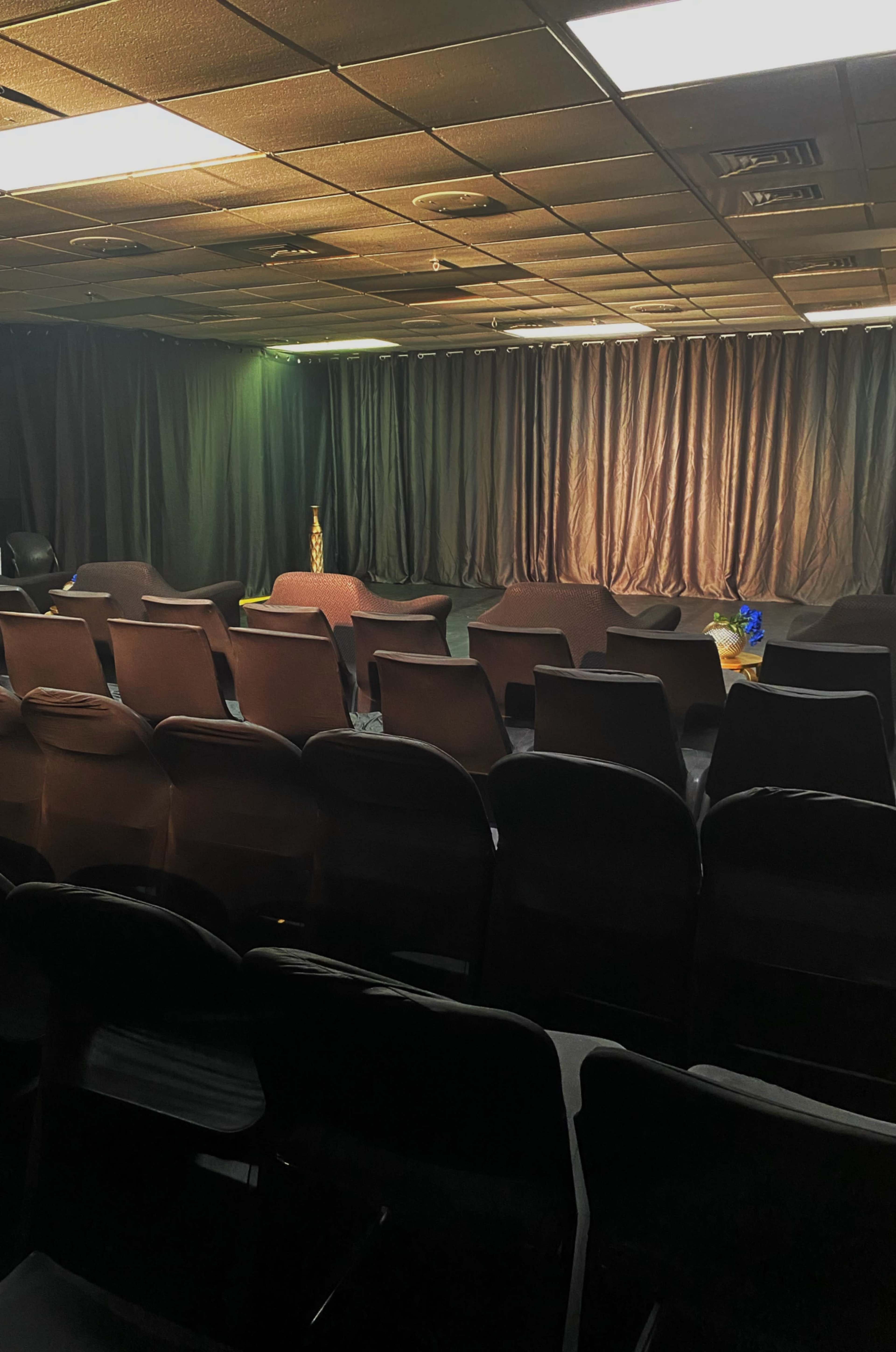 The image shows a dimly lit performance space with rows of black chairs facing a stage adorned with a backdrop of dark curtains.