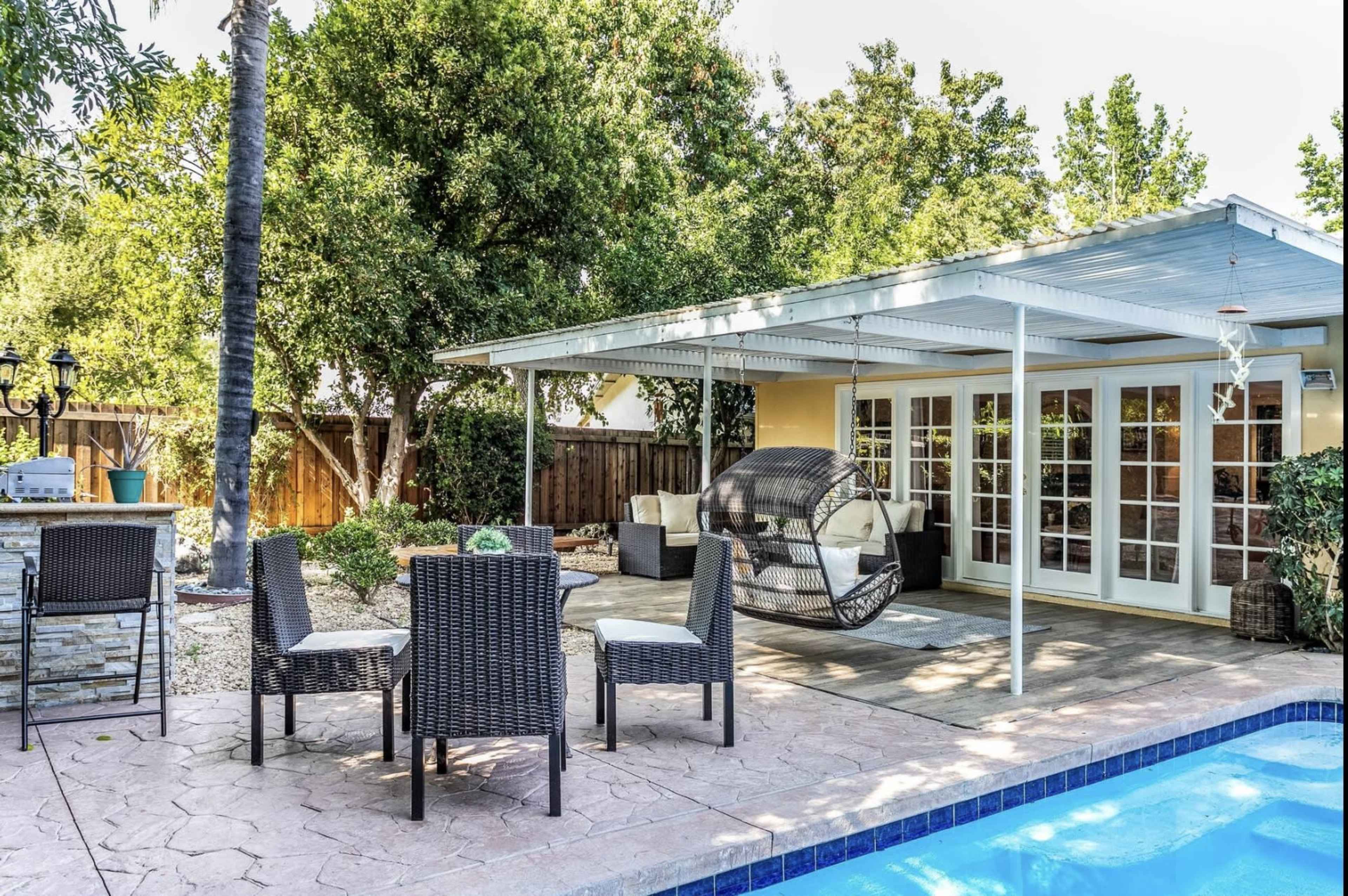 A backyard patio area with a hanging chair, a dining table set, and a swimming pool surrounded by greenery.