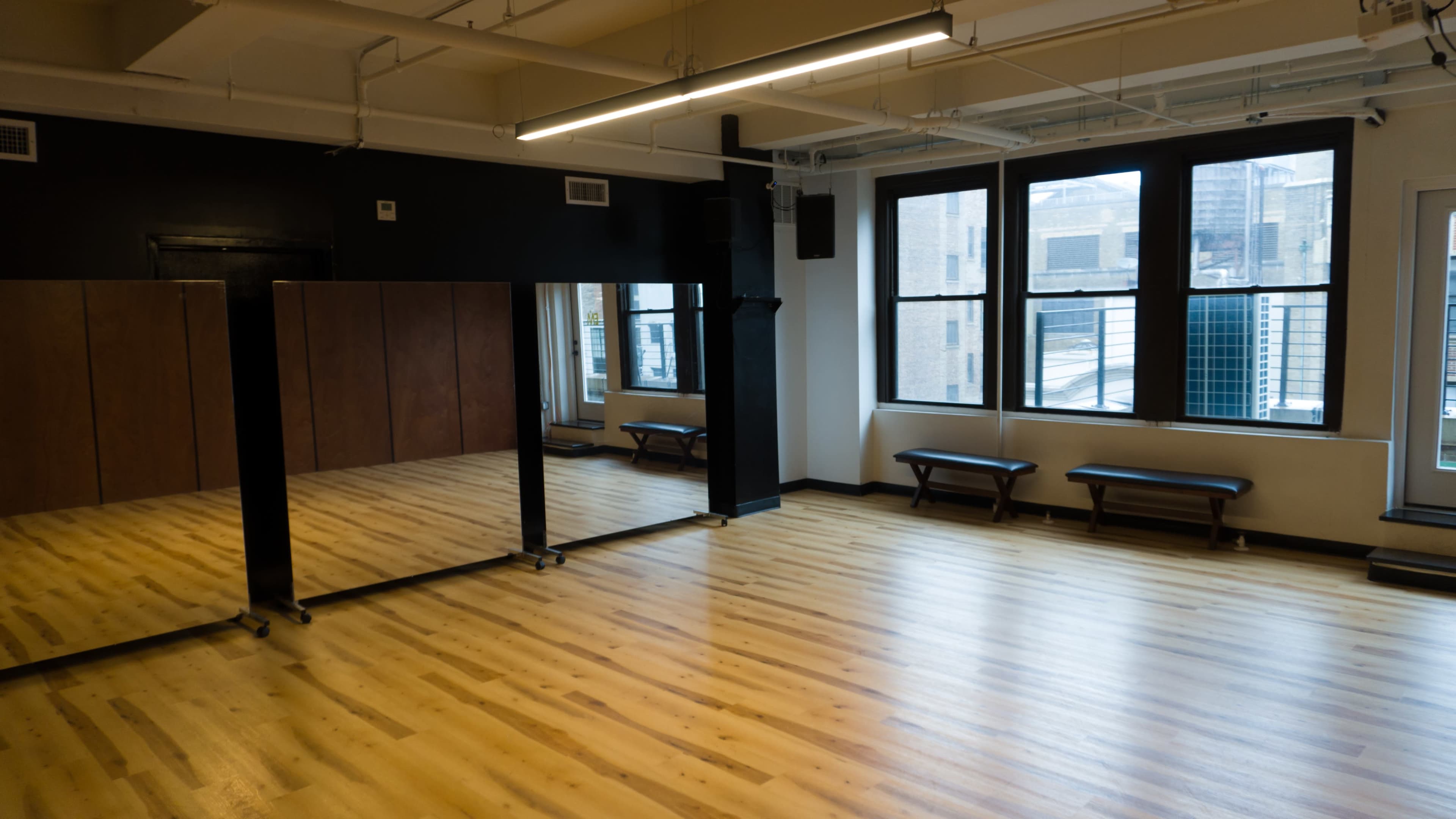The image shows a dance studio with hardwood floors, large mirrors on one wall, and benches along the opposite wall near large windows.