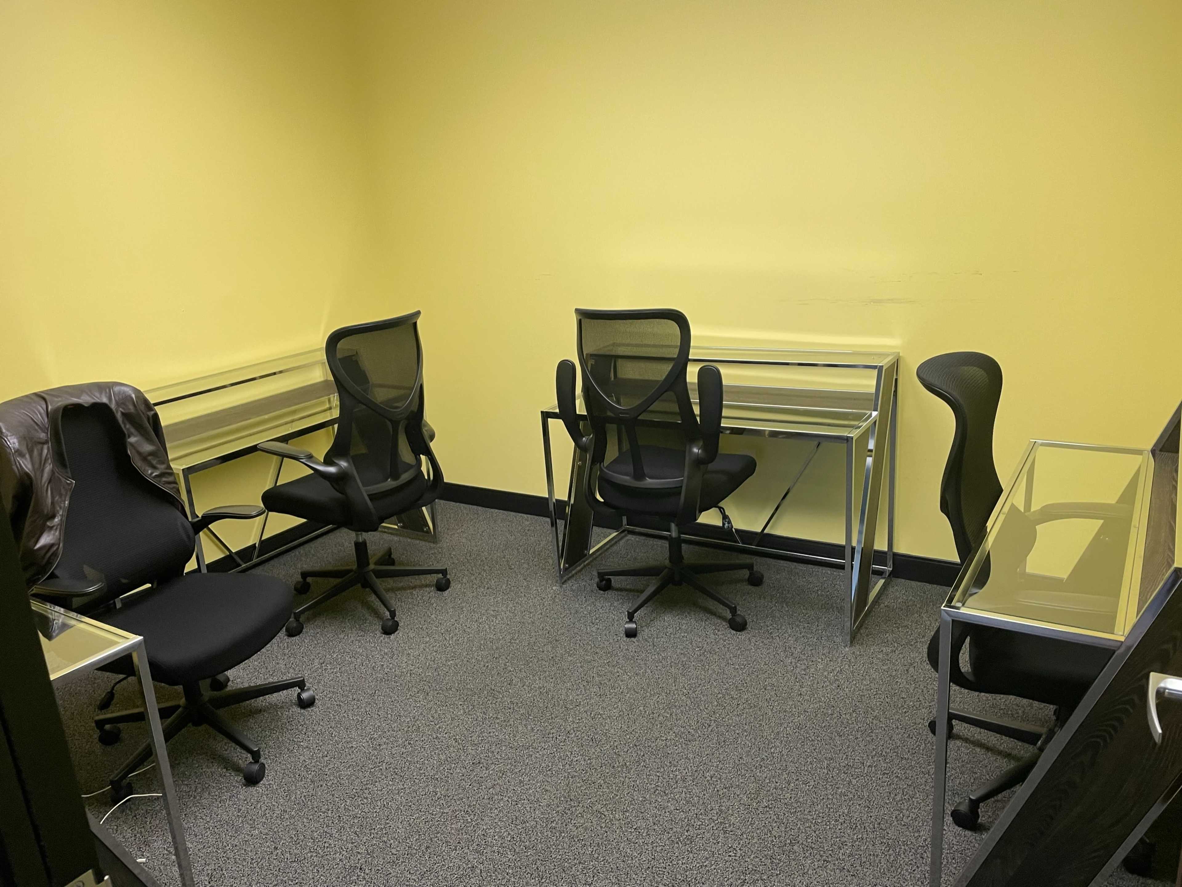 An empty room with yellow walls, containing several black office chairs and glass-top desks arranged in a circular layout.