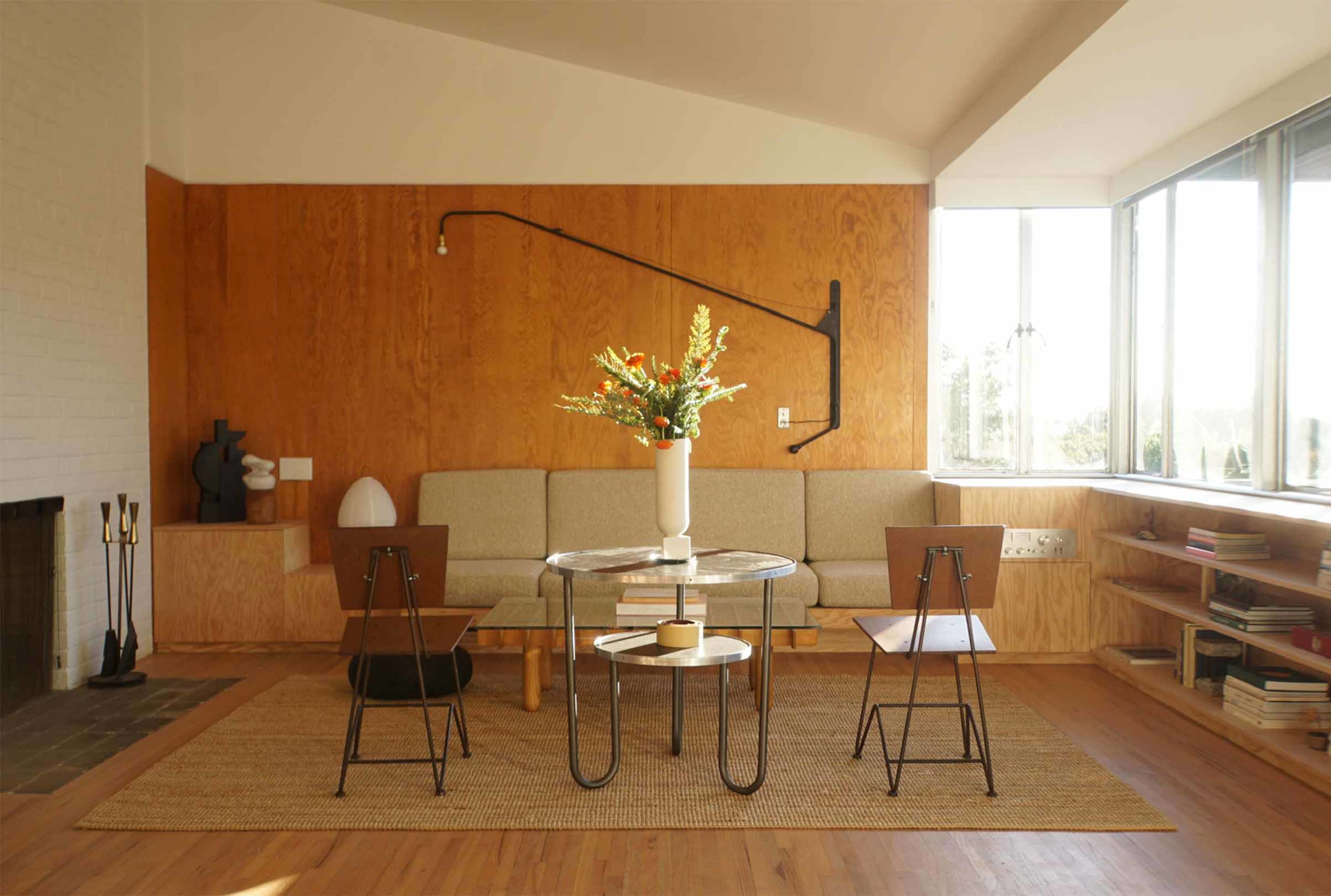 The image shows a modern living room with a wooden accent wall, a circular glass coffee table, two metal chairs, and a vase of flowers on the table.