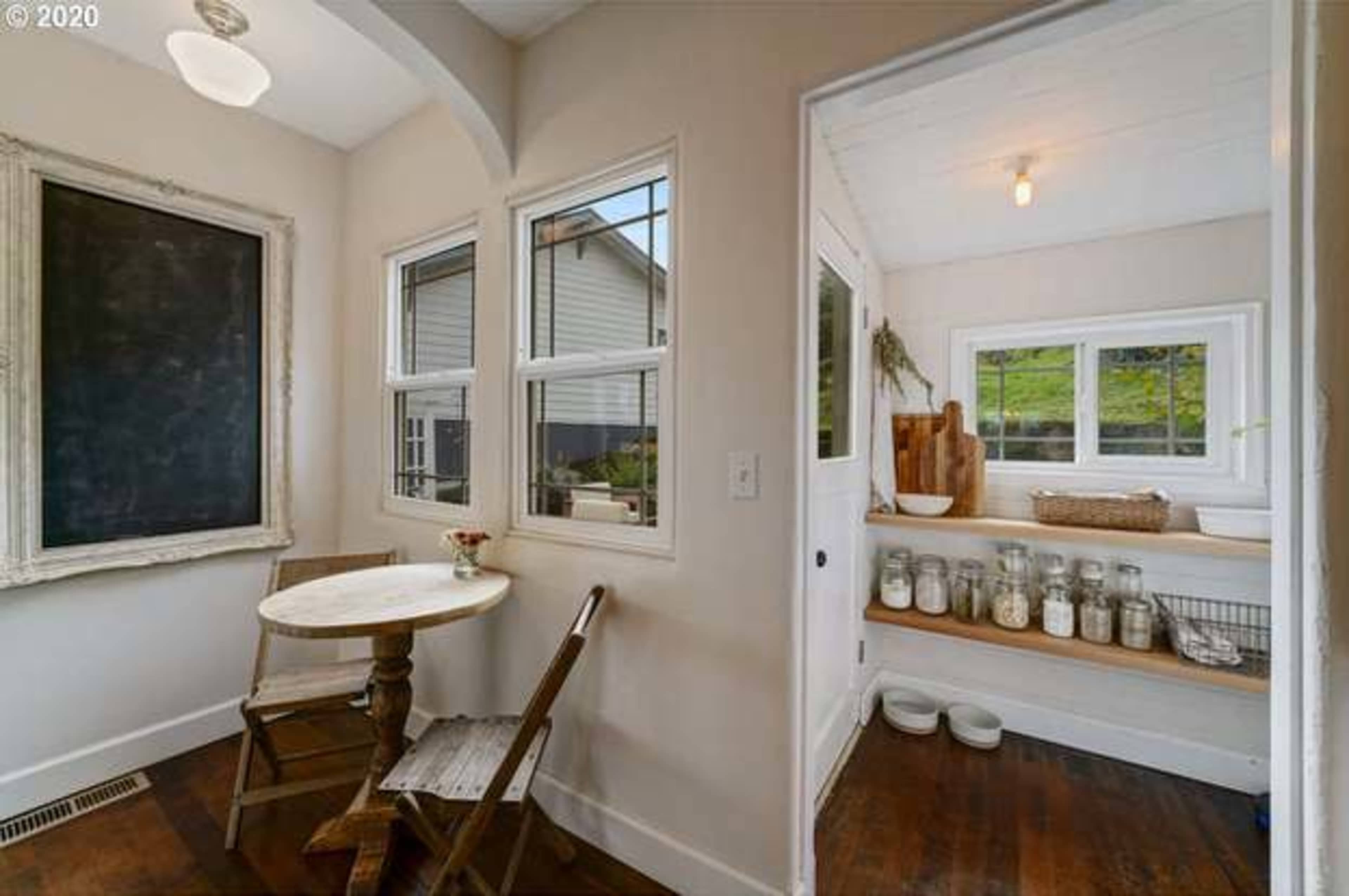 A small kitchen space features a round table with a single chair, a chalkboard on the wall, and a windowed nook displaying jars and a wooden cutting board.