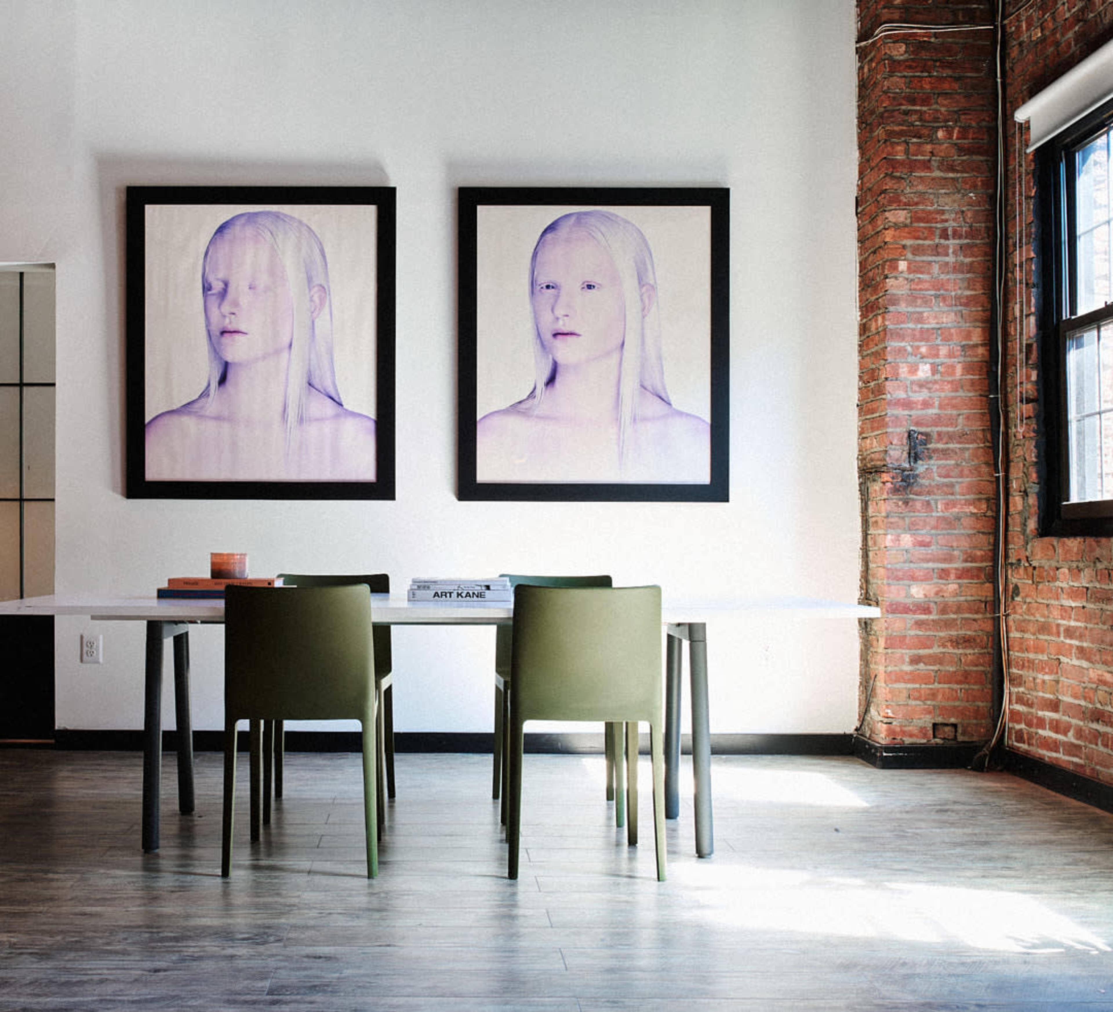 A modern dining area with a white table, green chairs, and two large framed portraits on the wall, set against exposed brick.