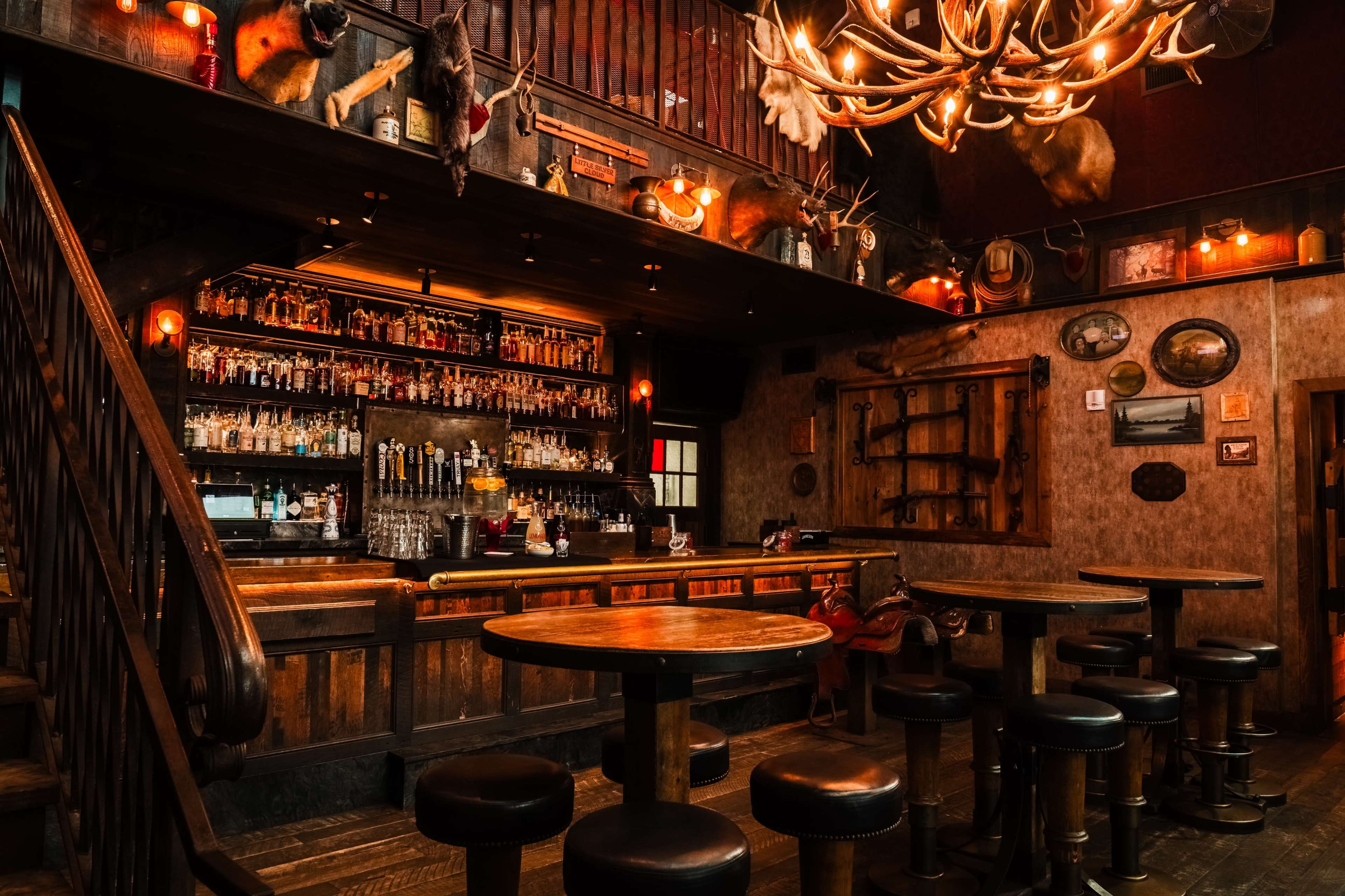 The image shows a rustic bar interior with wooden furnishings, a well-stocked back bar, and animal mounts decorating the walls.