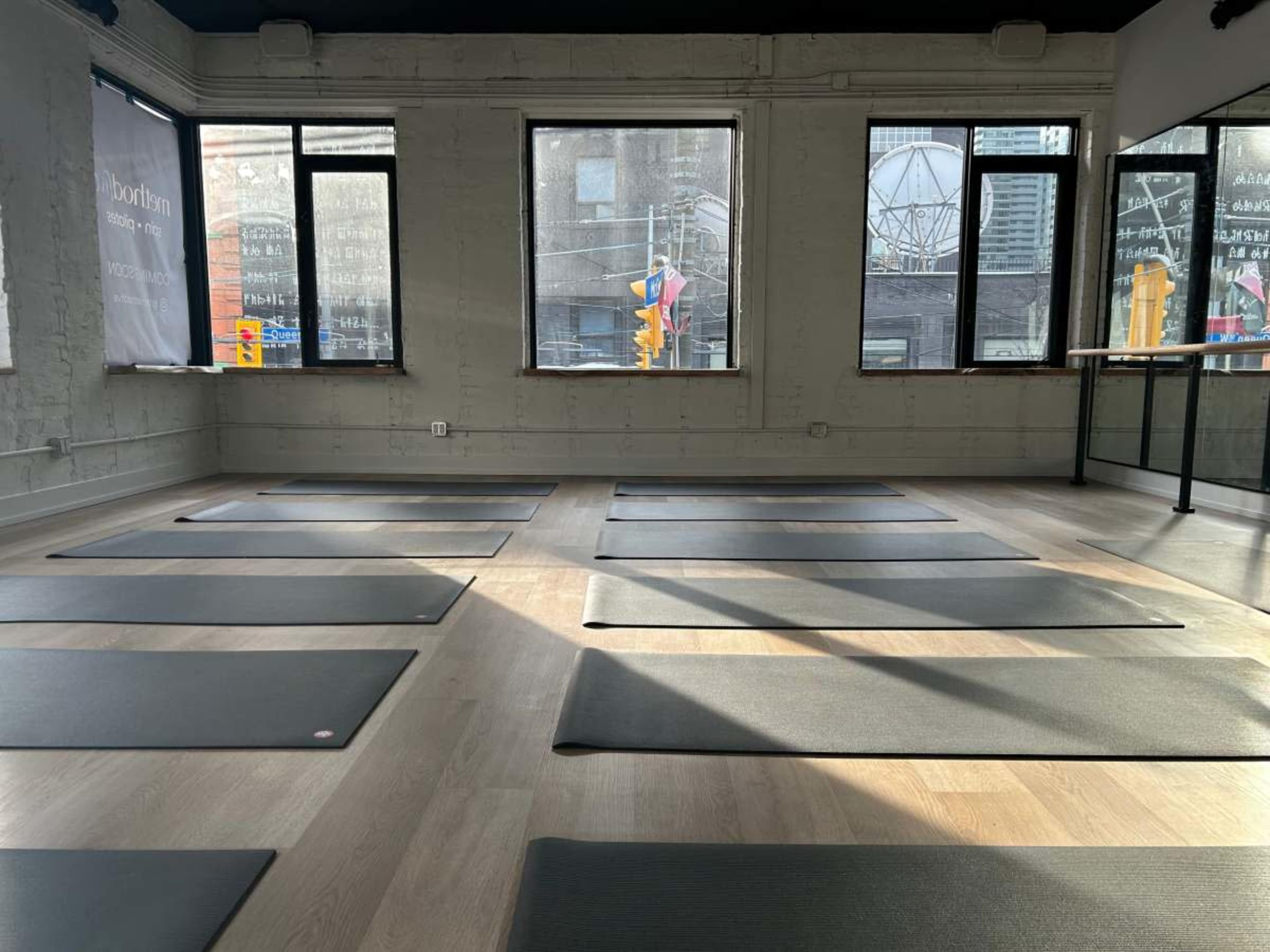 The image shows a yoga studio with several yoga mats arranged neatly on the wooden floor beneath large windows.