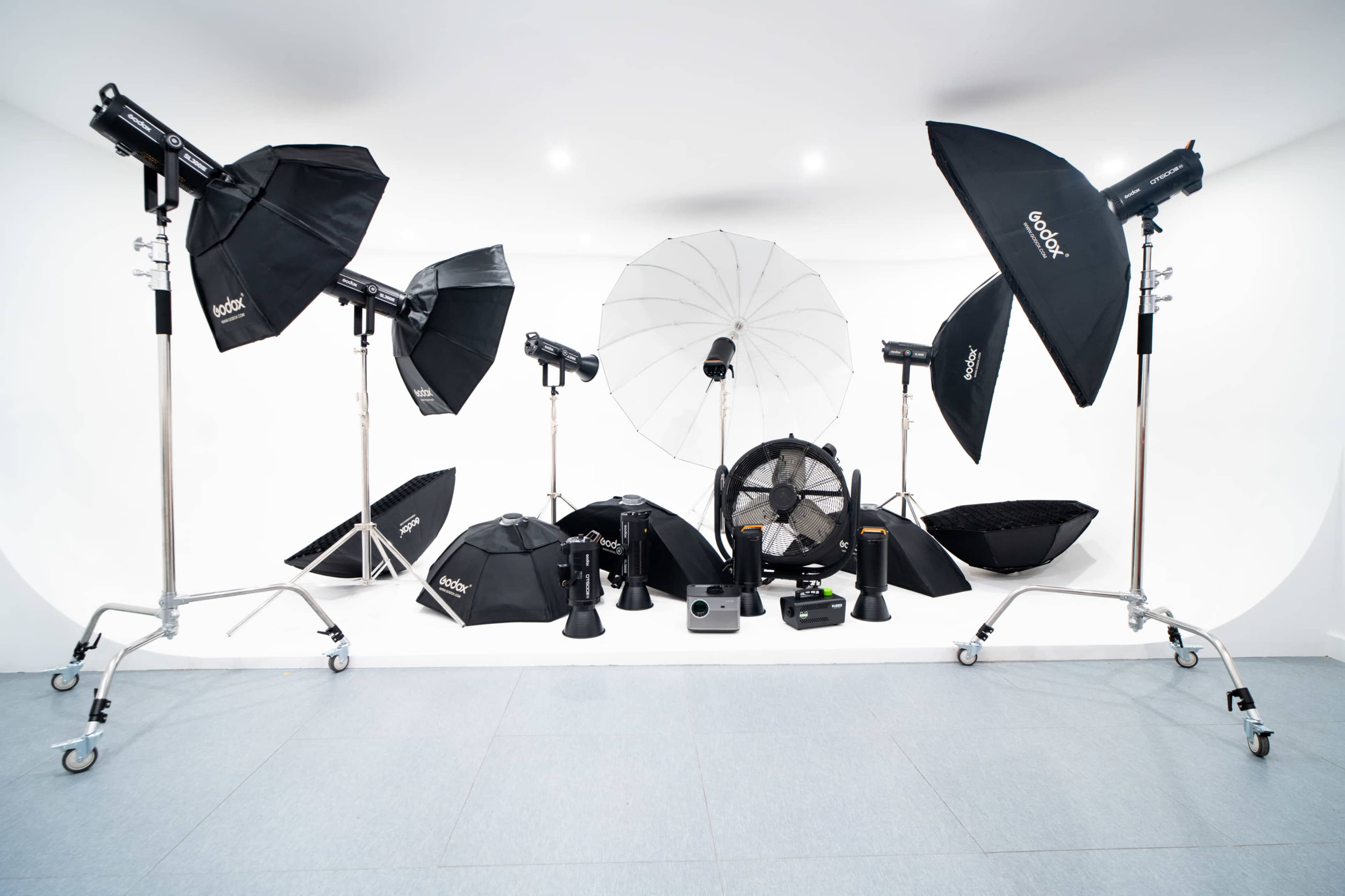 The image shows a photography studio setup featuring various lighting equipment, including softboxes, umbrellas, and light stands, arranged against a white backdrop.