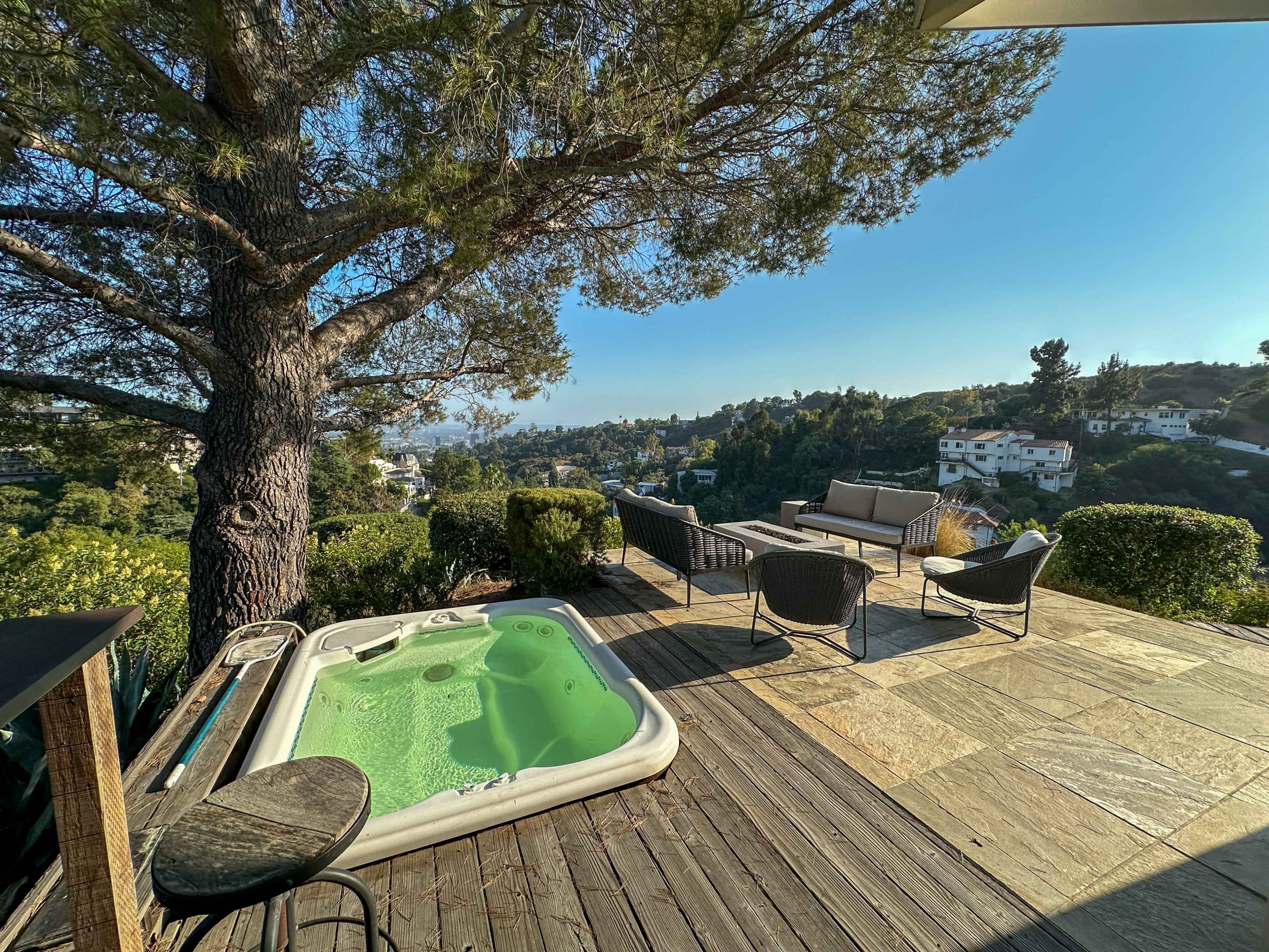 A small hot tub sits next to a seating area on a wooden deck overlooking a valley and hillside homes.