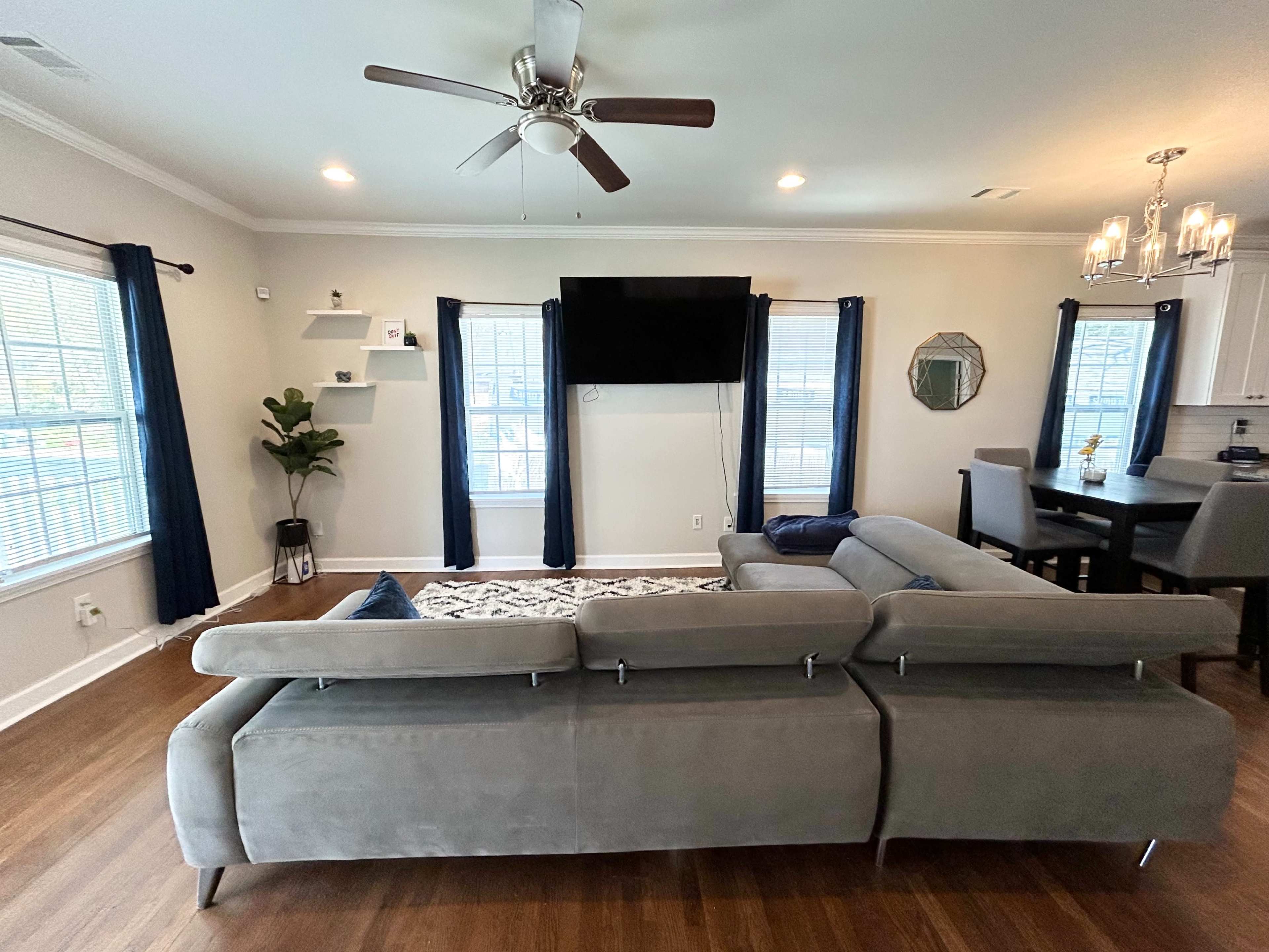 The image shows a modern living room with a sectional sofa, a large television mounted on the wall, and windows dressed with blue curtains.