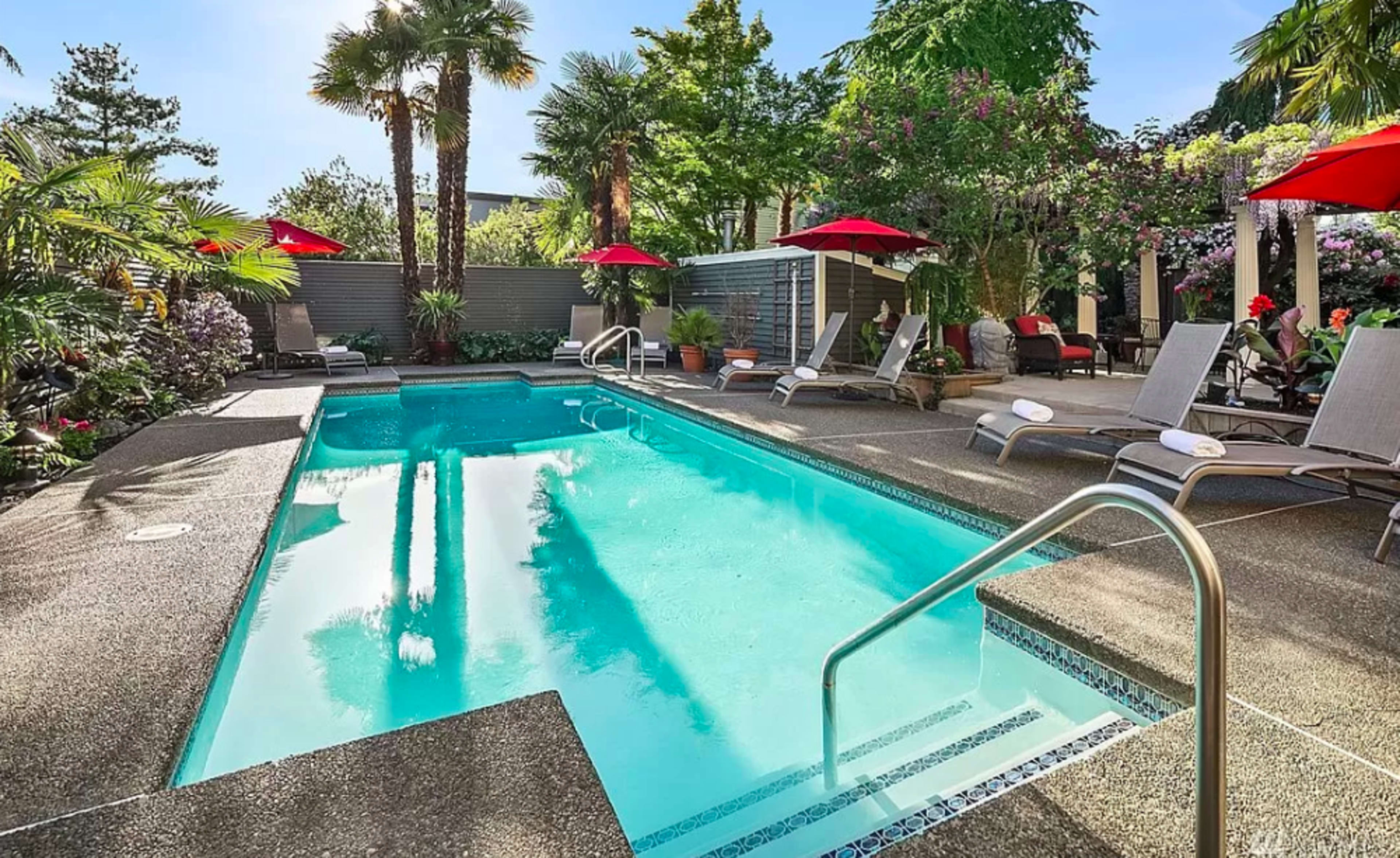 A swimming pool surrounded by palm trees and lounge chairs, with red umbrellas providing shade.